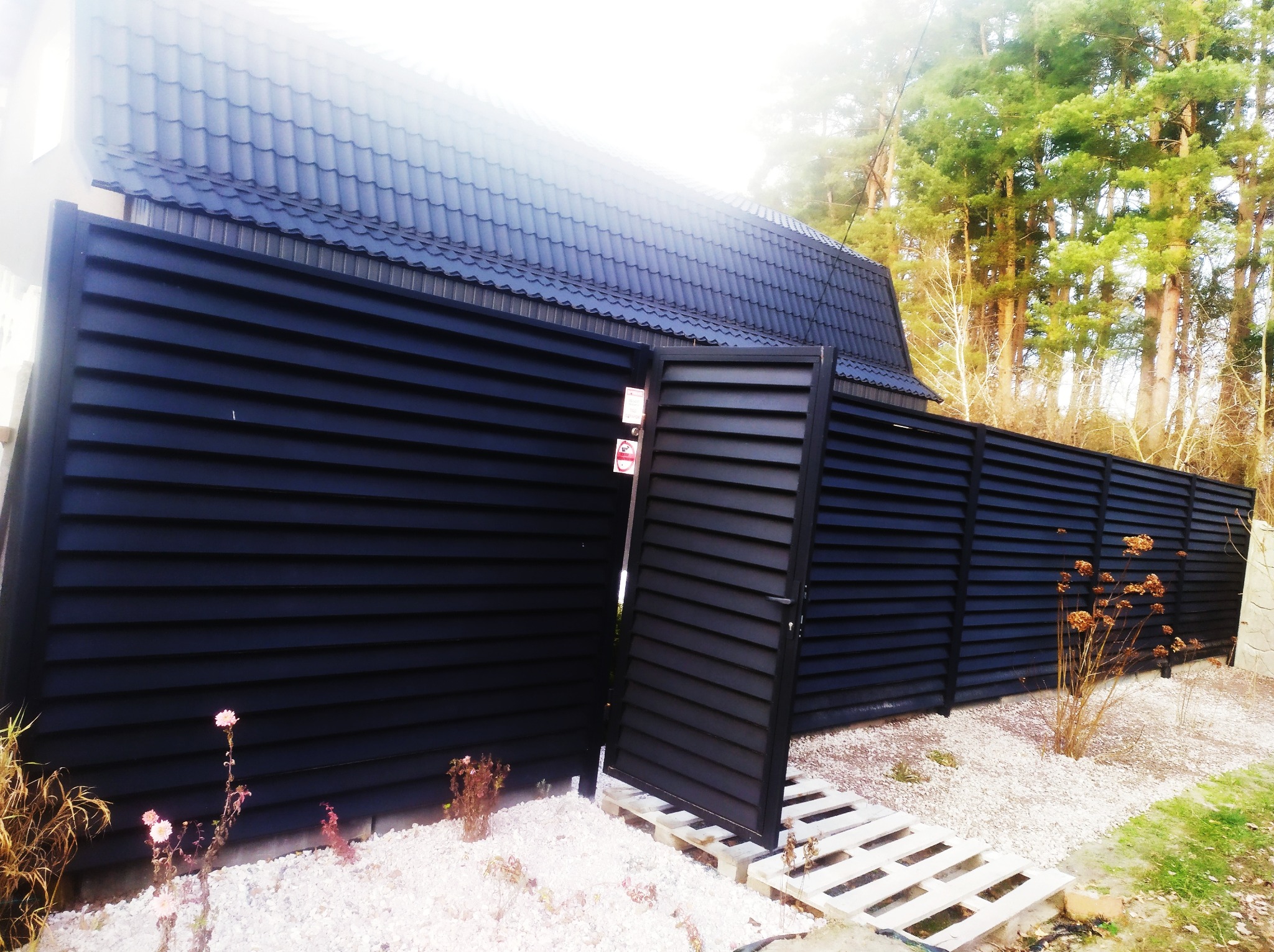 Nowoczesne, czarne ogrodzenie panelowe z otwartą furtką, ułożoną na palecie, na tle białego żwiru i domu z ciemnym dachem.