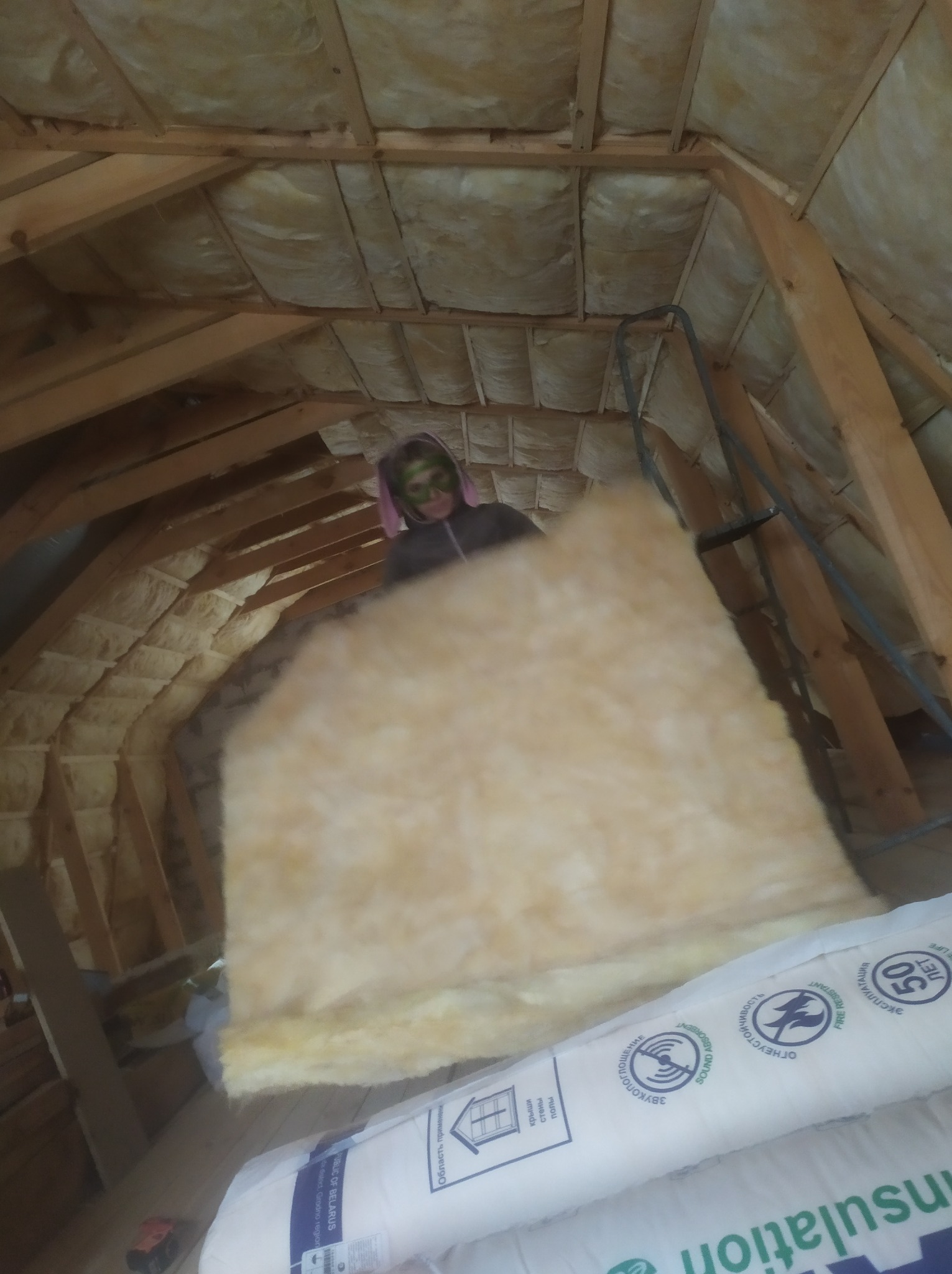 Osoba z maską na twarzy instaluje izolację z wełny mineralnej na poddaszu z drewnianą konstrukcją więźby dachowej, widoczna drabina i opakowania izolacji.