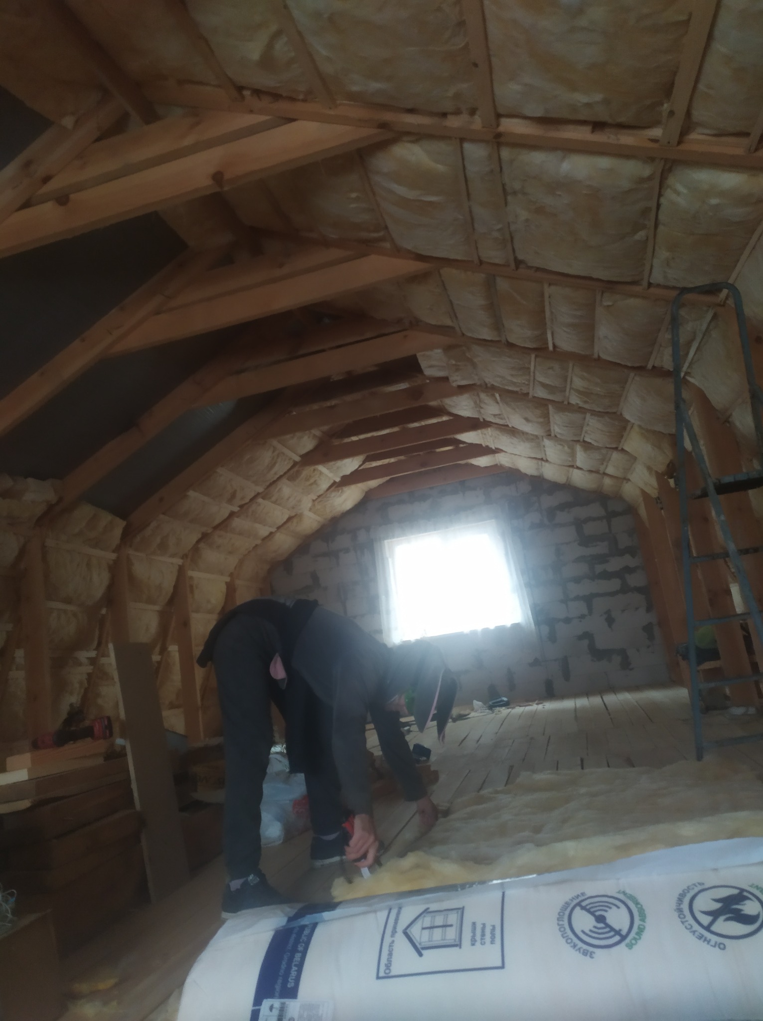 Pracownik w trakcie ocieplania poddasza wełną mineralną, widoczne drewniane krokwie, okno i częściowo wykończona ściana z cegły.