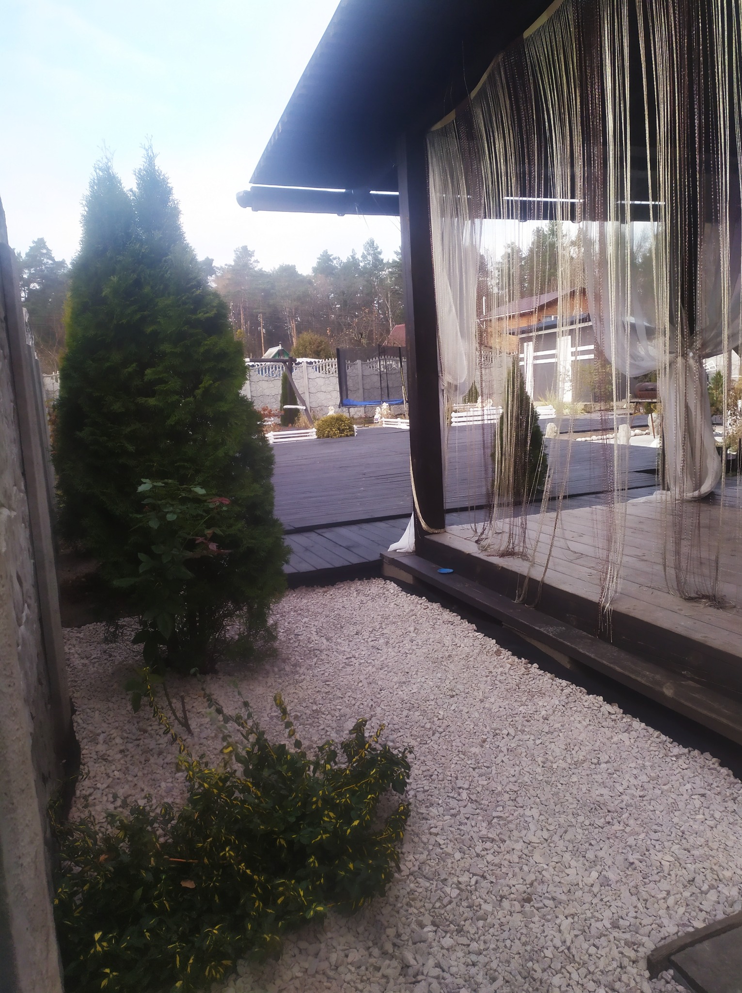 Zadaszony taras z drewnianą podłogą i dekoracyjnymi zasłonami, otoczony białymi kamieniami i zielenią, z widokiem na ogród i dom w tle.