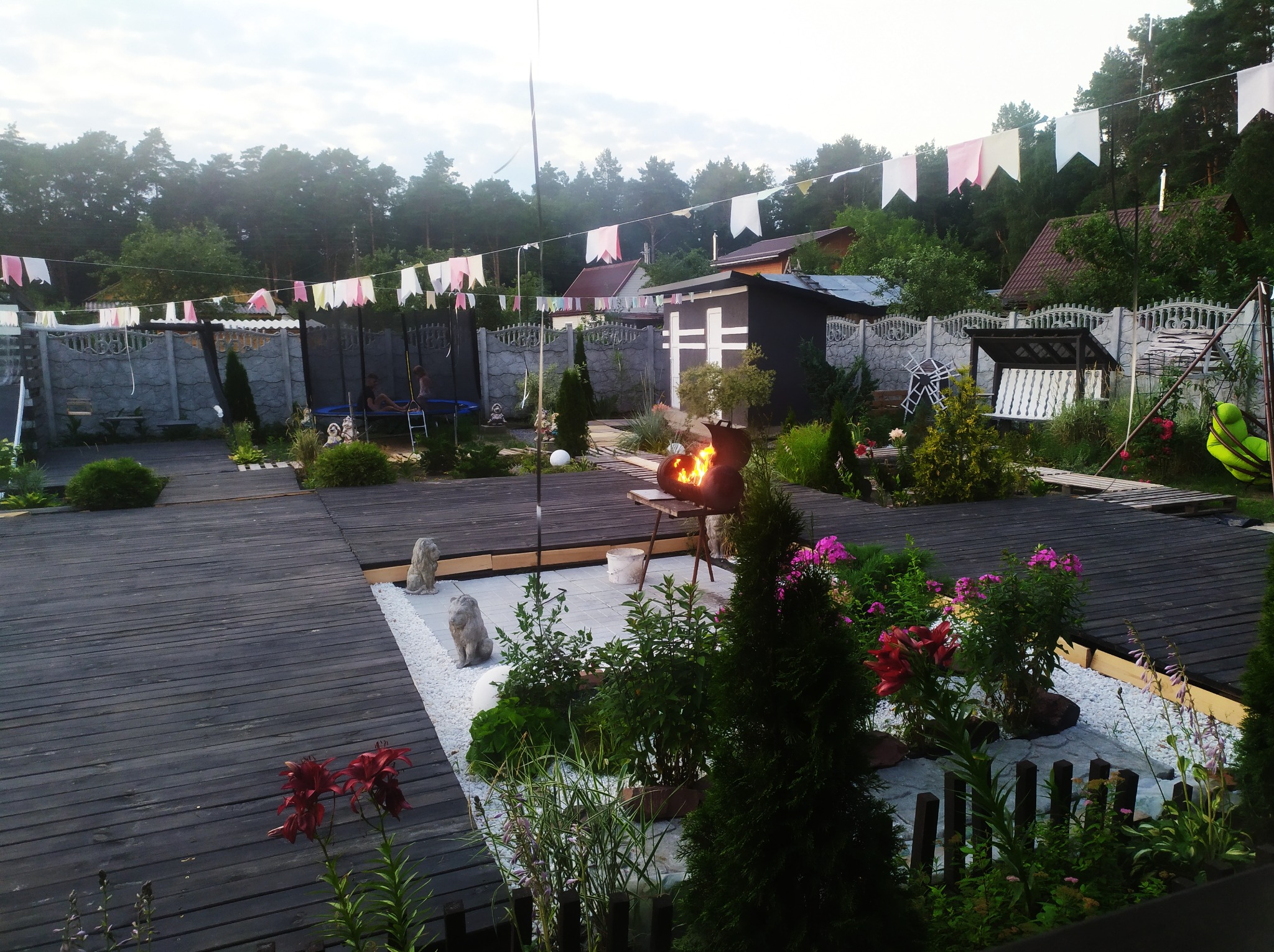 Szeroki, drewniany taras z kwiatami, dekoracyjnymi kamieniami i rzeźbami zwierząt, grill z płomieniami w tle, ozdobne flagi rozwieszone nad przestrzenią, widoczne elementy ogrodzenia i zabudowy...
