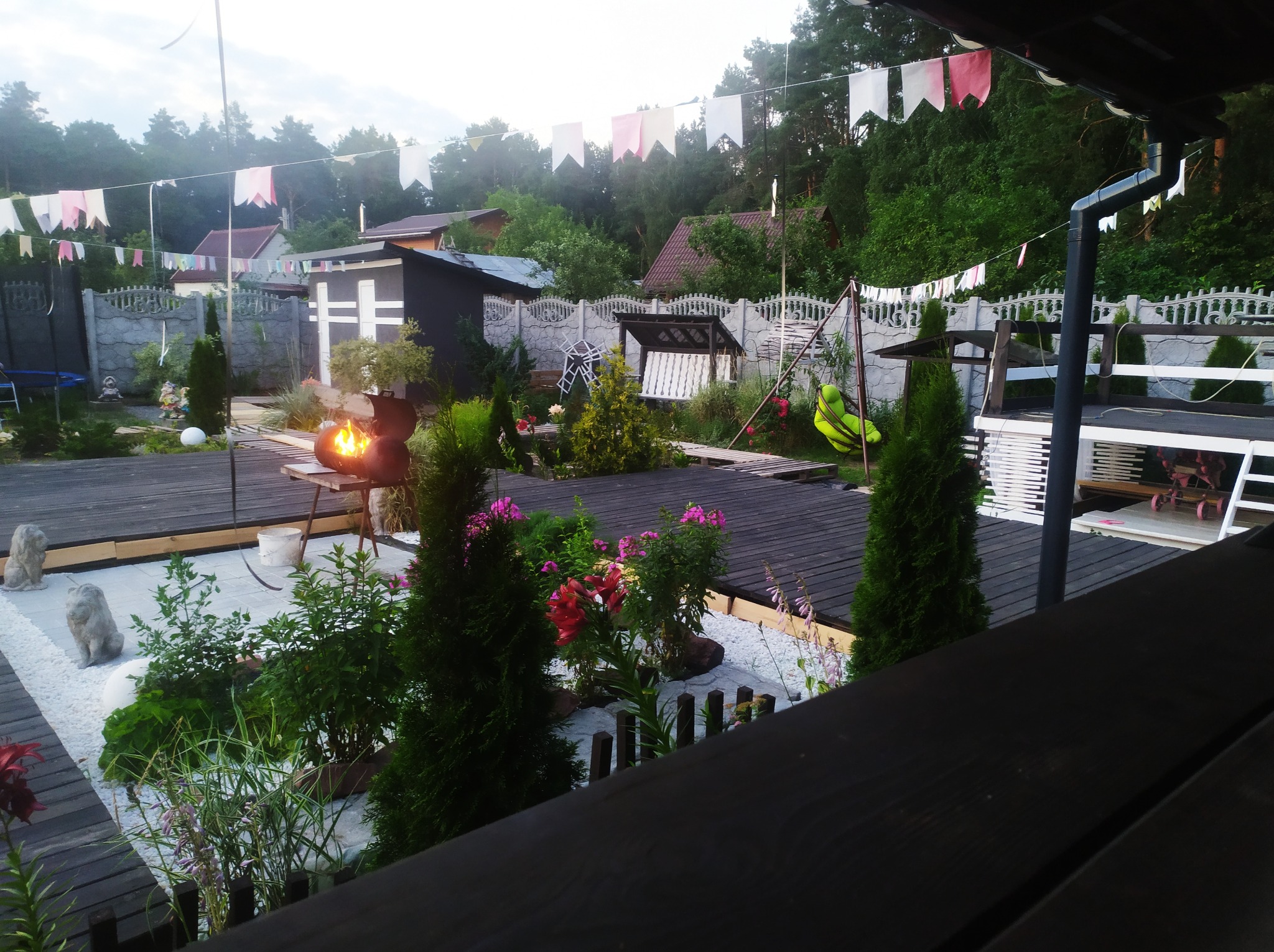 Urokliwy ogród z drewnianym tarasem, białymi kamieniami, dekoracyjnymi figurkami lwów, i płonącym grillem, ozdobiony girlandami z chorągiewek, otoczony zielenią drzew.