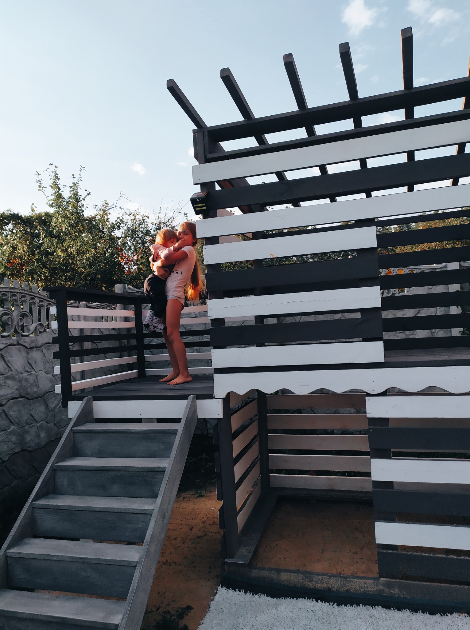 Dzieci na drewnianej konstrukcji z elementami pomalowanymi na czarno i biało, przypominającej domek lub plac zabaw, z schodami i ogrodzeniem z imitacji kamienia w tle.