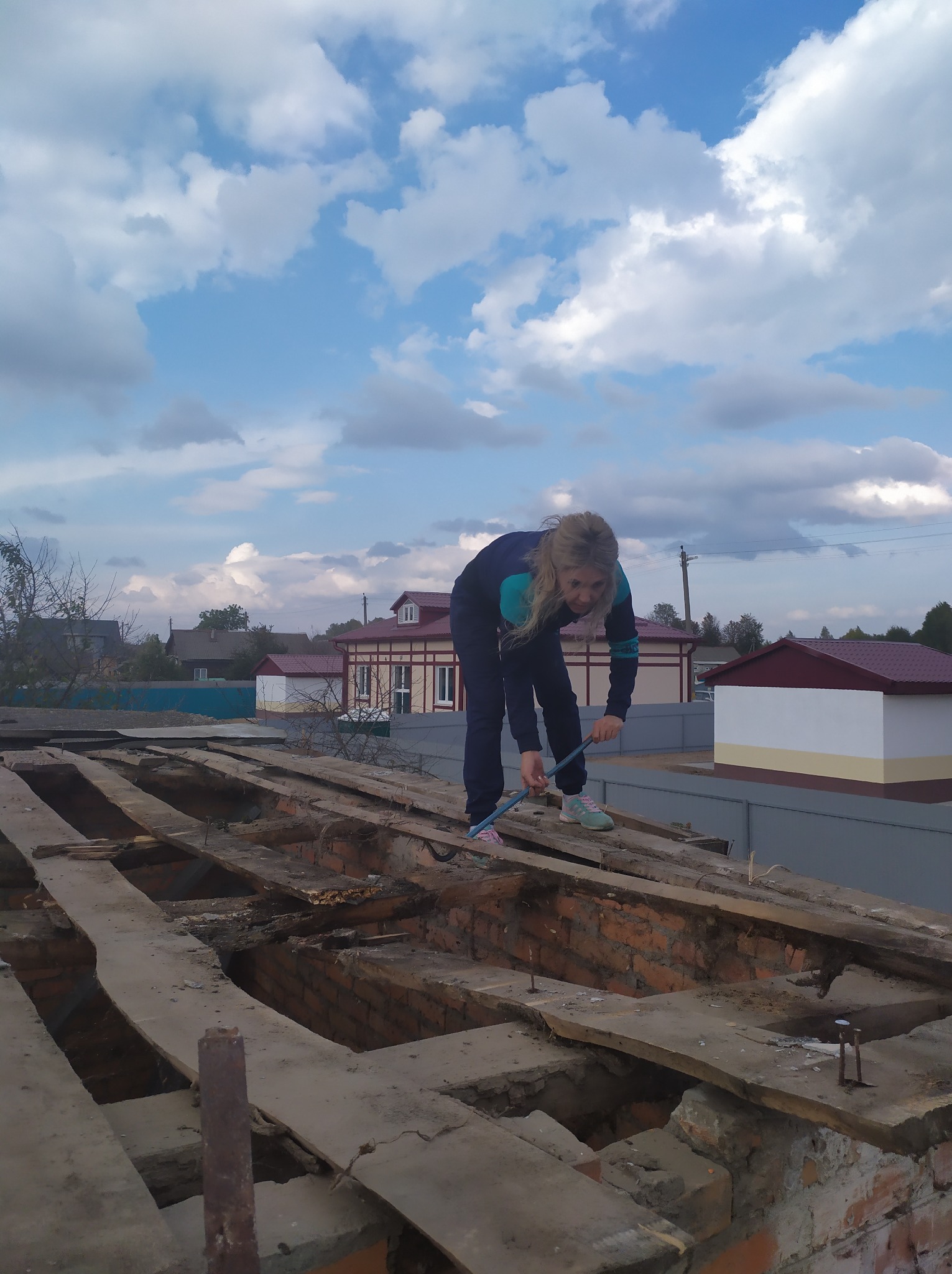 Kobieta w dresie czyści szczotką konstrukcję dachu z drewnianymi belkami i ceglanymi przestrzeniami, na tle błękitnego nieba z chmurami i zabudowań.