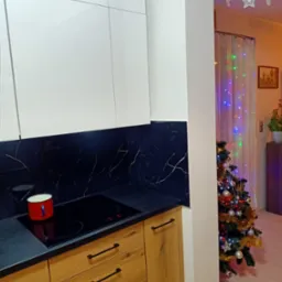 Nowoczesna kuchnia z białymi szafkami górnymi i drewnianymi dolnymi, czarny blat z marmurowym wzorem, płyta indukcyjna z czerwonym garnkiem, w tle choinka i świąteczne dekoracje.