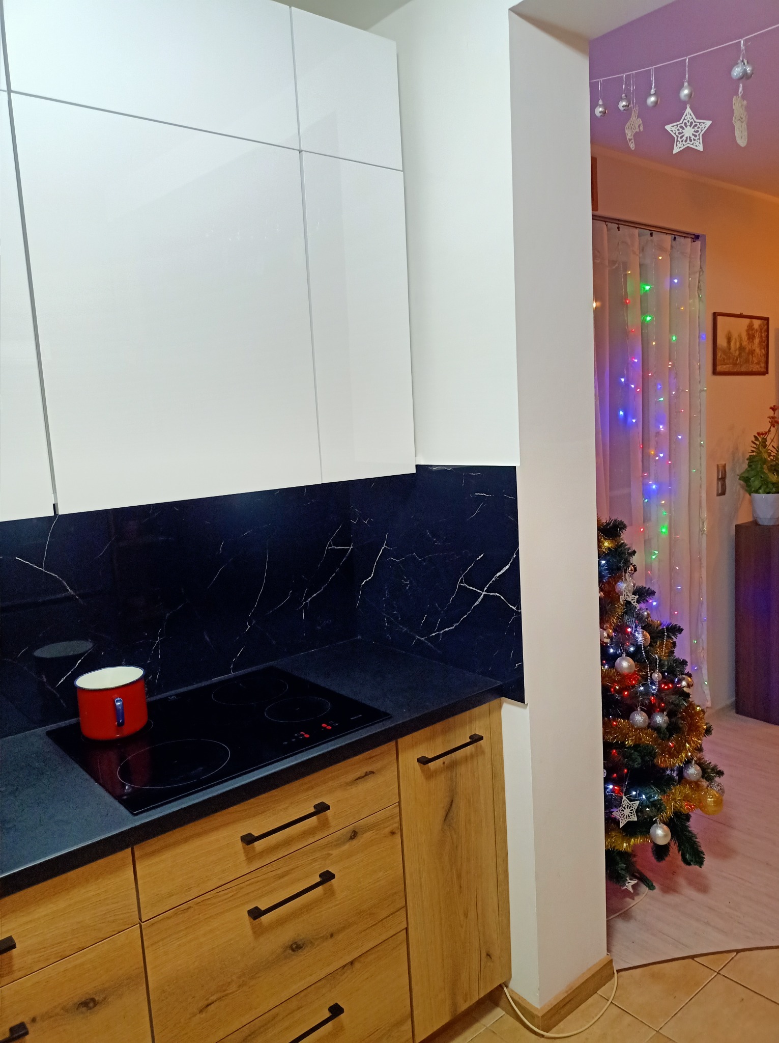 Nowoczesna kuchnia z białymi szafkami górnymi i drewnianymi dolnymi, czarny blat z marmurowym wzorem, płyta indukcyjna z czerwonym garnkiem, w tle choinka i świąteczne dekoracje.