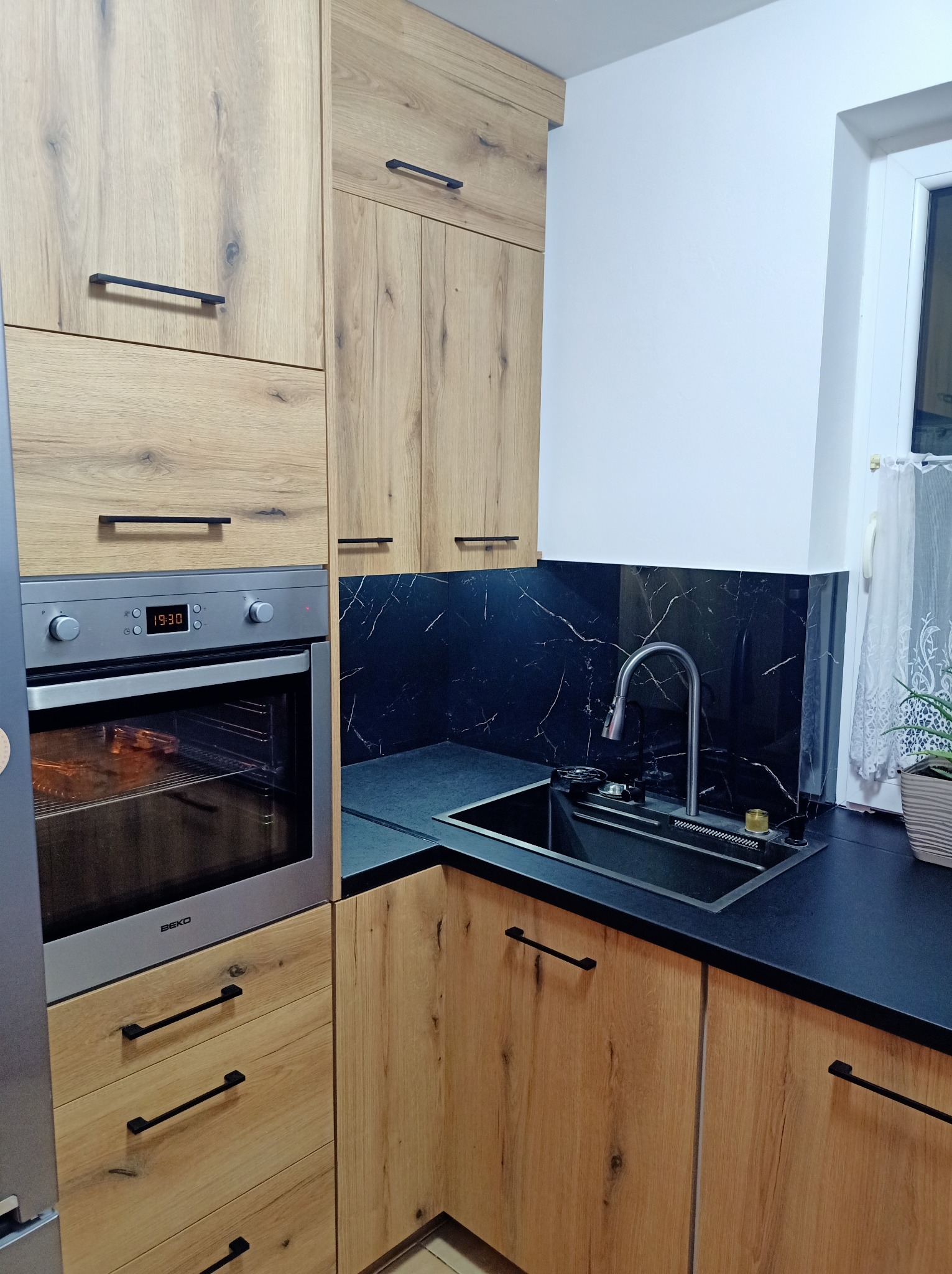 Fragment nowoczesnej kuchni z drewnianymi frontami szafek, czarnym blatem i panelem nad zlewem imitującym marmur, widoczny piekarnik marki Beko.