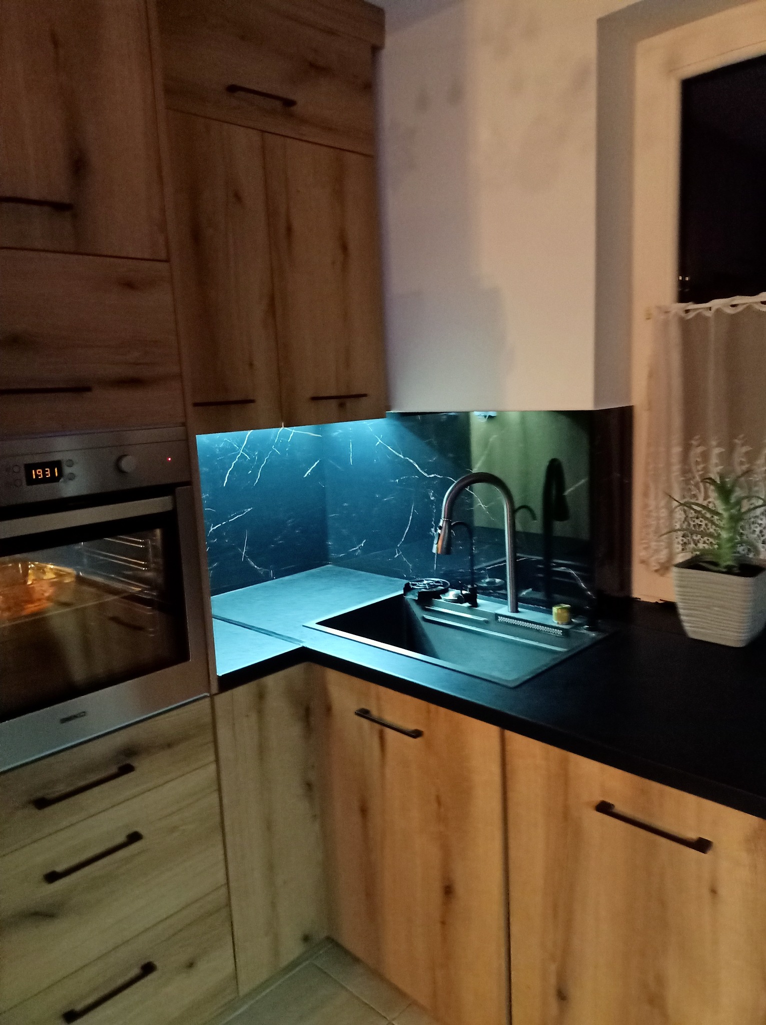 Fragment drewnianej zabudowy kuchennej z czarnym blatem, zlewozmywakiem i blatem nad zlewem z motywem marmuru, podświetlonym od dołu. W tle okno z firanką i doniczką z rośliną.
