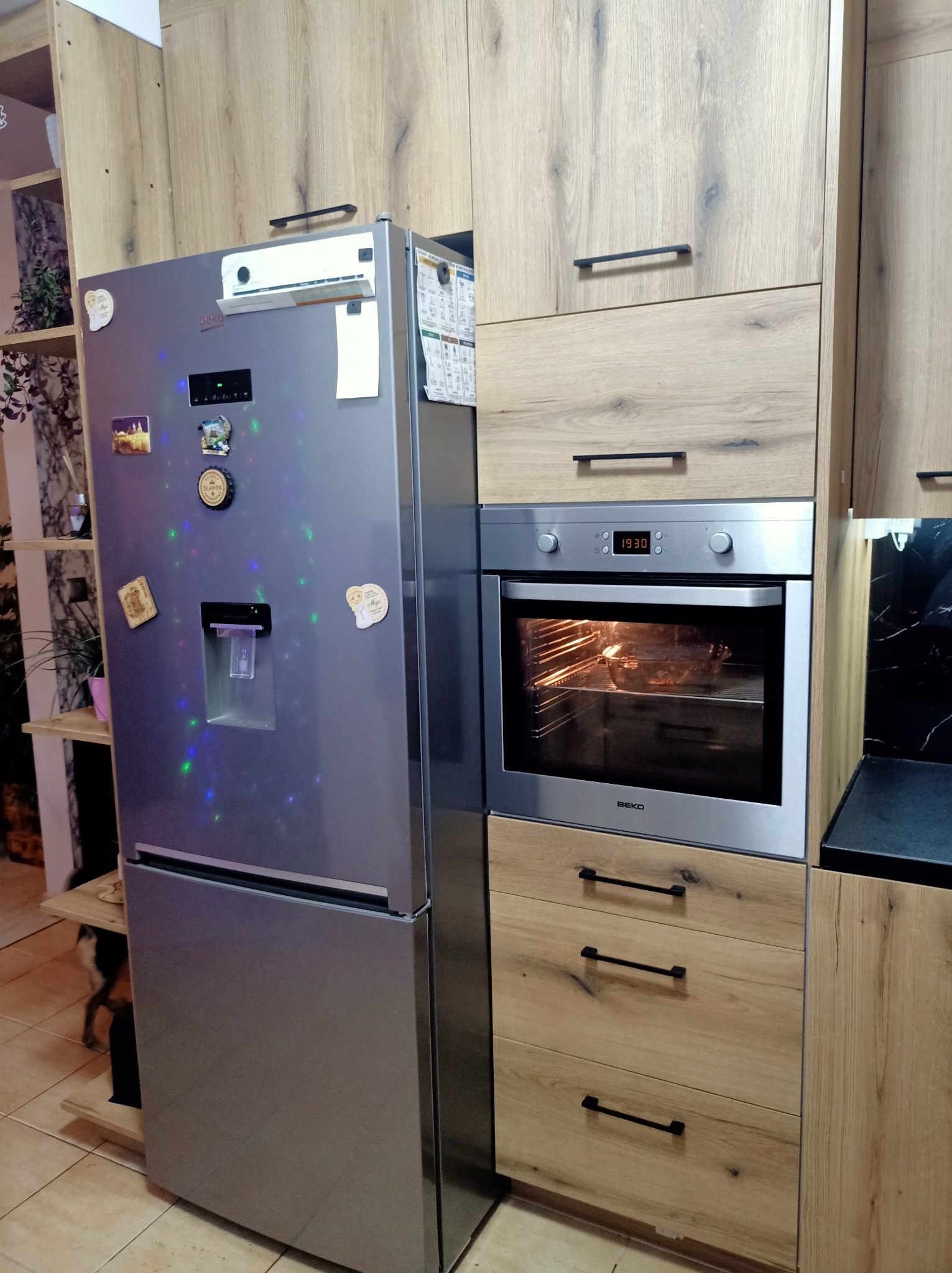 Szara lodówka z magnesami i dystrybutorem wody obok zabudowanej piekarnika Beko w kuchni z drewnianymi szafkami z czarnymi uchwytami.