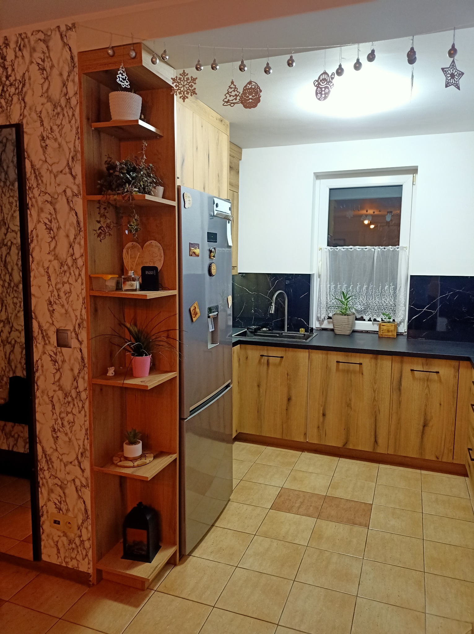 Jasna kuchnia z drewnianymi frontami, czarnym blatem i marmurkową ścianą, ozdobiona świątecznymi dekoracjami i roślinami doniczkowymi, z widoczną lodówką i otwartym regałem.