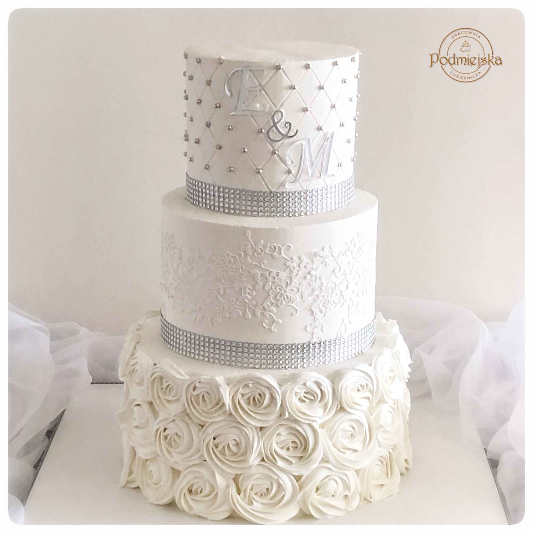 Trzypiętrowy tort weselny w odcieniach bieli, zdobiony kremowymi różami na dole, koronkowym wzorem na środkowym piętrze i kryształkami oraz inicjałami 'E & M' na górnym piętrze, z logo cukierni...