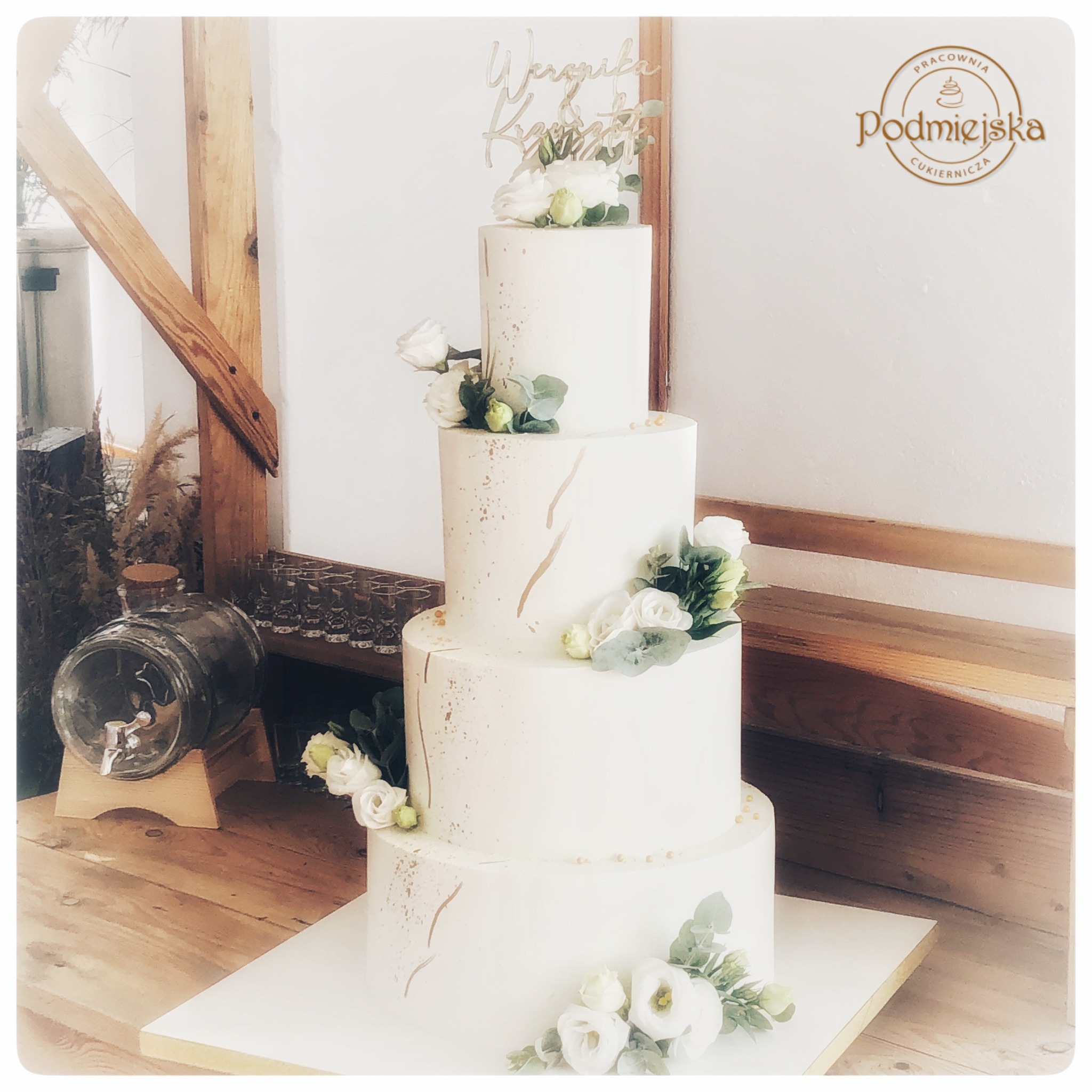 Czteropiętrowy tort weselny w stylu rustykalnym, zdobiony białymi kwiatami i złotymi akcentami, z drewnianym topperem z imionami, prezentowany na tle drewnianej konstrukcji i szklanego dystrybutora...