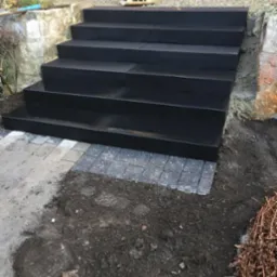 Nowe, czarne, kamienne schody prowadzące do domu, z widoczną ubijarką gruntu na górze i fragmentem kostki brukowej u podstawy, otoczone ziemią i roślinnością.