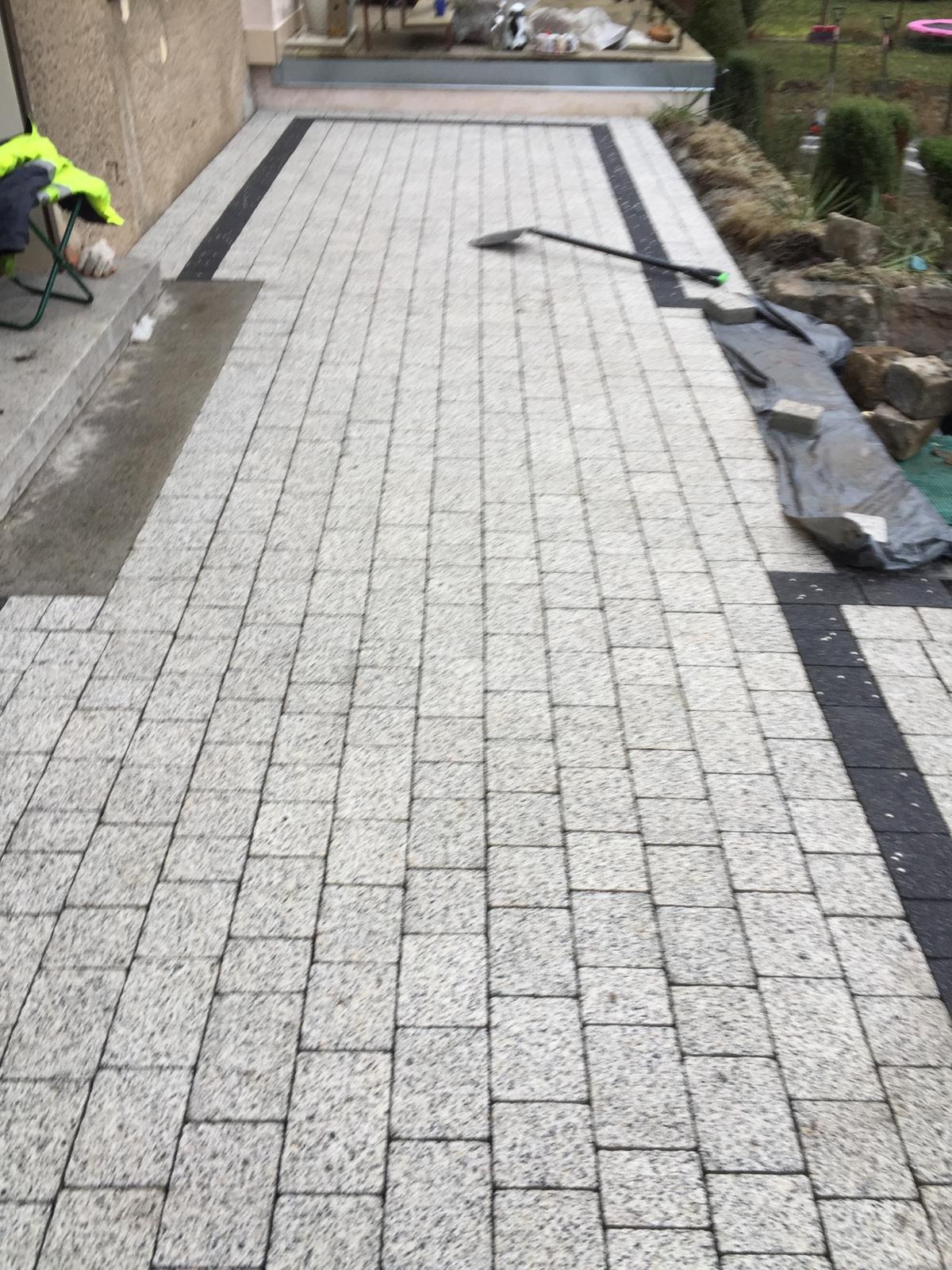 Ułożona ścieżka z jasnoszarej kostki brukowej z ciemnymi pasami po bokach, widoczna łopata i fragmenty ogrodu w tle.