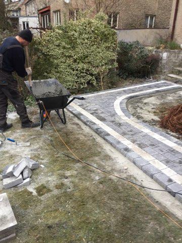 Pracownik budowlany w czarnym ubraniu z łopatą przy taczce pełnej zaprawy, obok nowo ułożony chodnik z kostki brukowej w szaro-białej kolorystyce na tle ogrodu i budynku.