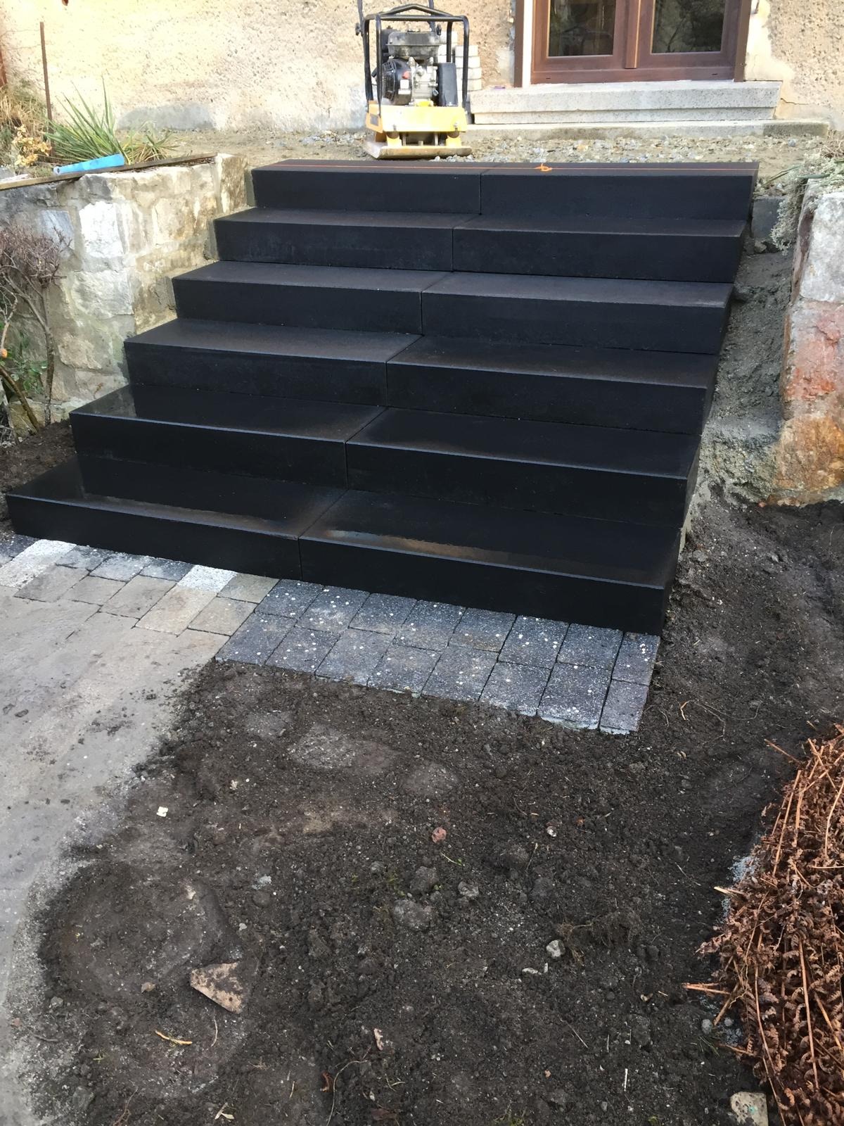 Nowe, czarne, kamienne schody prowadzące do domu, z widoczną ubijarką gruntu na górze i fragmentem kostki brukowej u podstawy, otoczone ziemią i roślinnością.