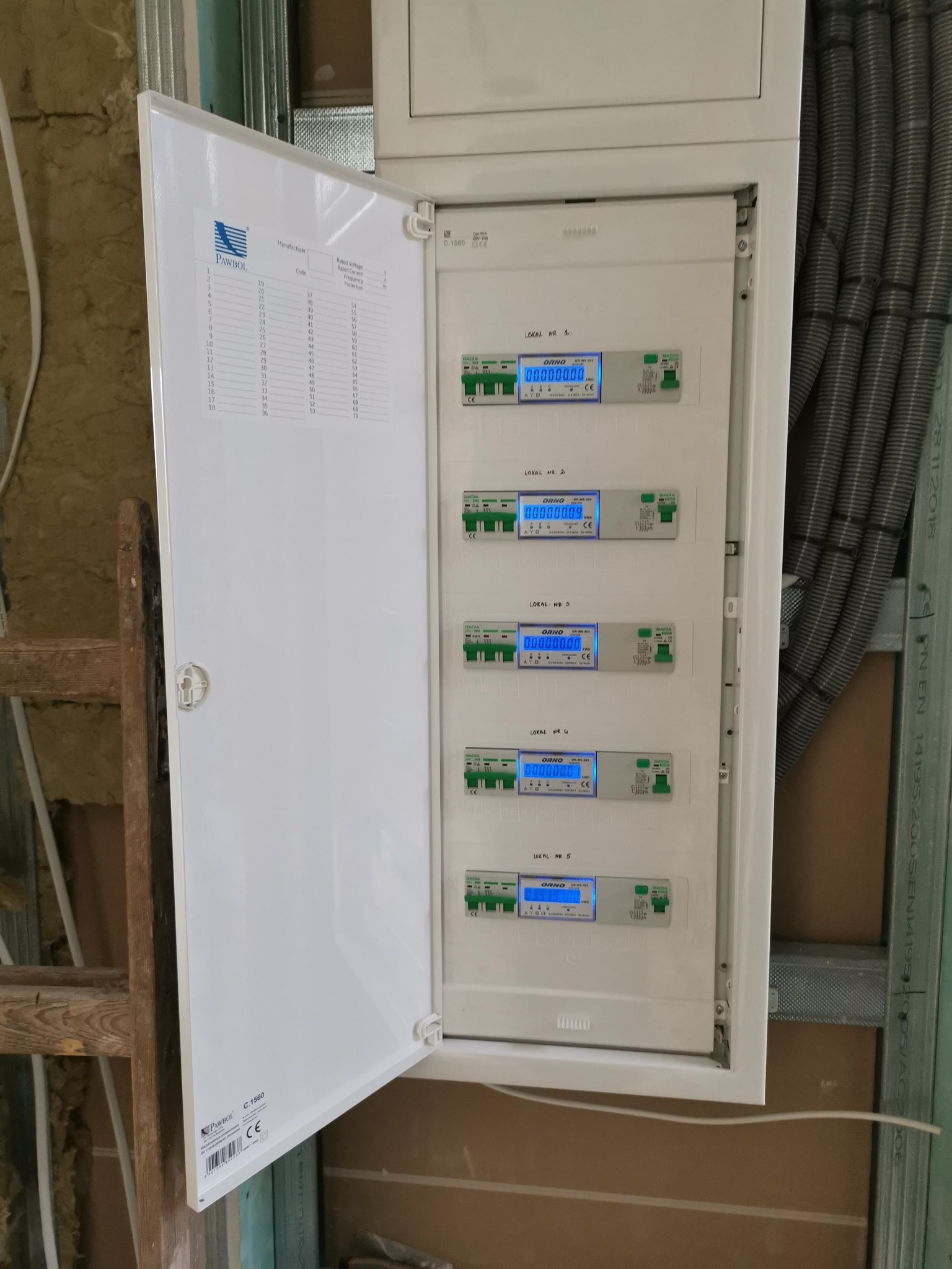 Otwarta skrzynka elektryczna z licznikami energii w surowym wnętrzu, widoczne przewody w peszlu i konstrukcja ścian działowych.