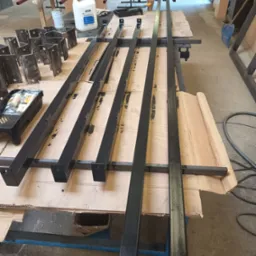 Przygotowane do montażu czarne, metalowe elementy konstrukcyjne, prawdopodobnie fragmenty balustrady lub ogrodzenia, leżą na stole warsztatowym w pomieszczeniu produkcyjnym. W tle widoczne inne...