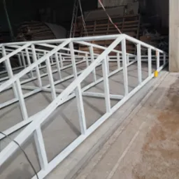 Biała, metalowa konstrukcja szkieletowa, prawdopodobnie brama lub ogrodzenie, leżąca na betonowej posadzce w hali produkcyjnej, widoczne ślady spawania i niedoskonałości powłoki malarskiej.