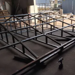 Konstrukcja szkieletowa z metalowych profili w hali produkcyjnej, widoczna wiertarka akumulatorowa, poziomica i fragmenty materiałów.