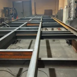 Konstrukcja stalowa z profili zamkniętych w warsztacie, widoczne spawy i przewody spawalnicze na podłodze, w tle wózek widłowy.