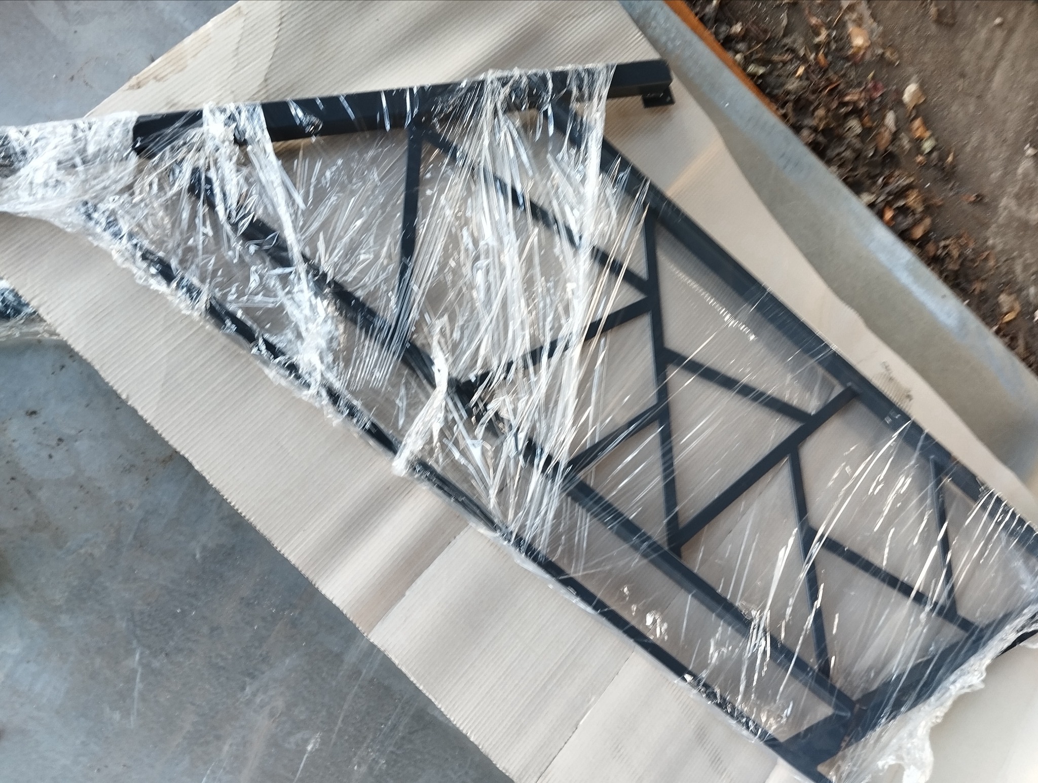 Część metalowej konstrukcji, prawdopodobnie fragment balustrady lub ogrodzenia, zabezpieczona folią stretch, leżąca na tekturze falistej.