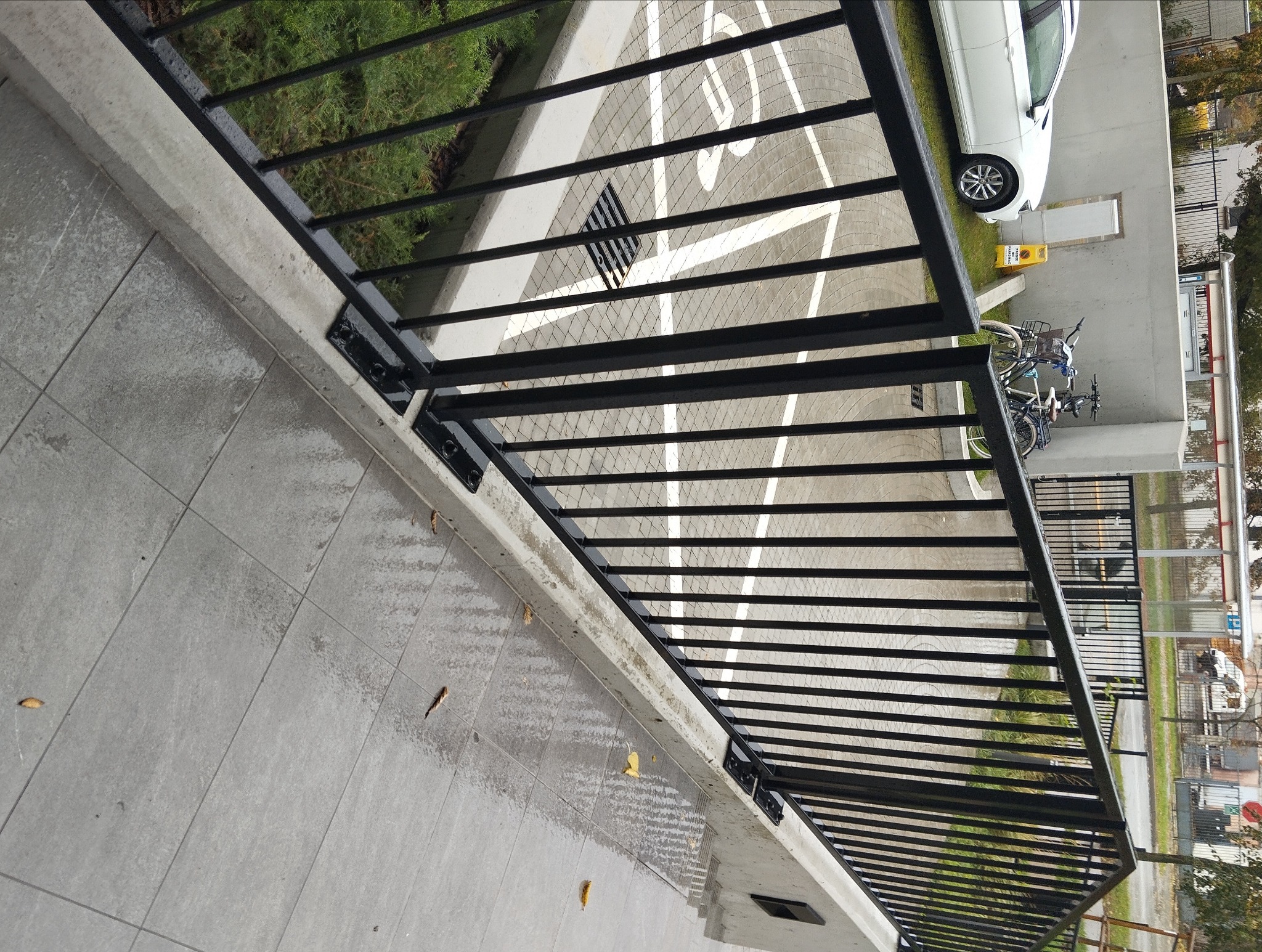 Czarna, metalowa balustrada balkonowa z widokiem na parking z białymi liniami, samochodem i rowerami w tle.