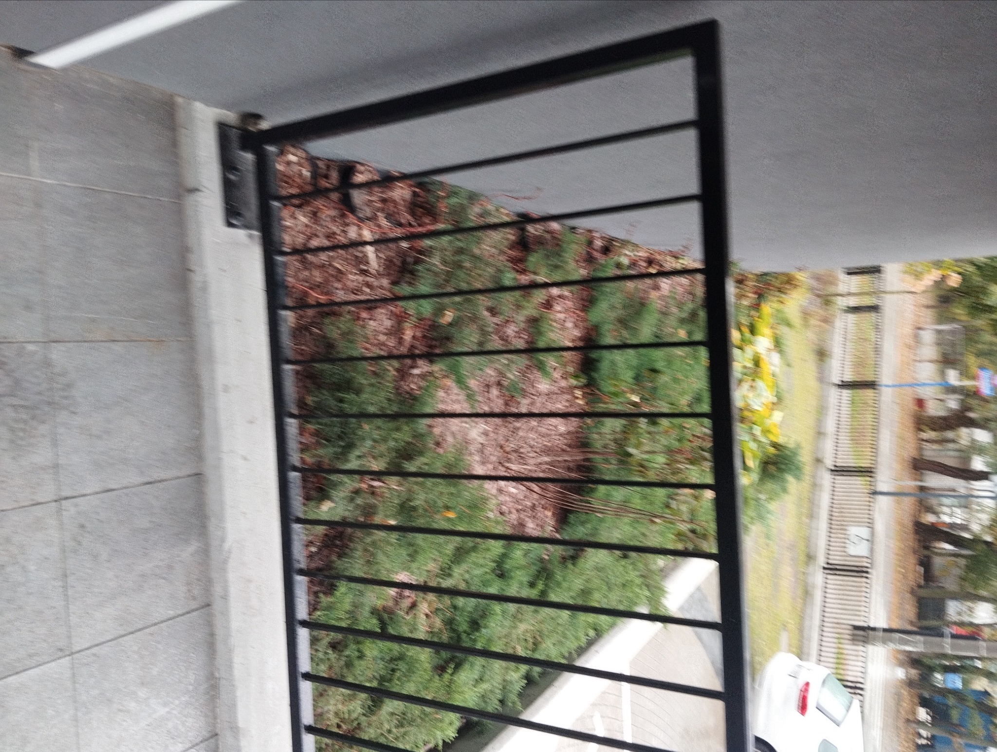 Częściowy widok czarnej, metalowej balustrady balkonowej z poziomymi prętami, zamocowanej do betonowej ściany, z widokiem na zieleń i fragment ulicy w tle.