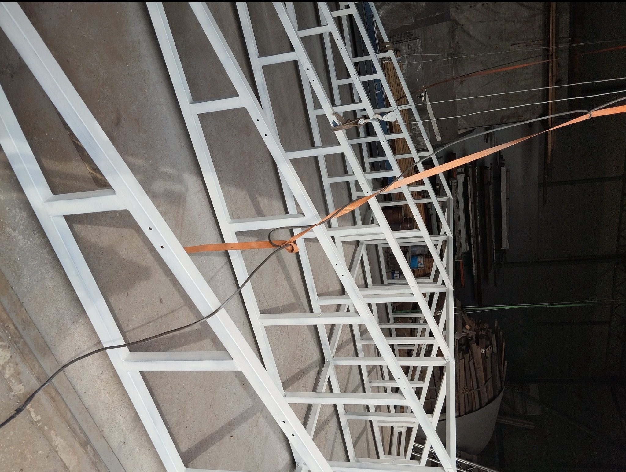 Biała, metalowa konstrukcja spawana z profili, zabezpieczona pasami transportowymi, widok z dołu, w tle ściana warsztatu.