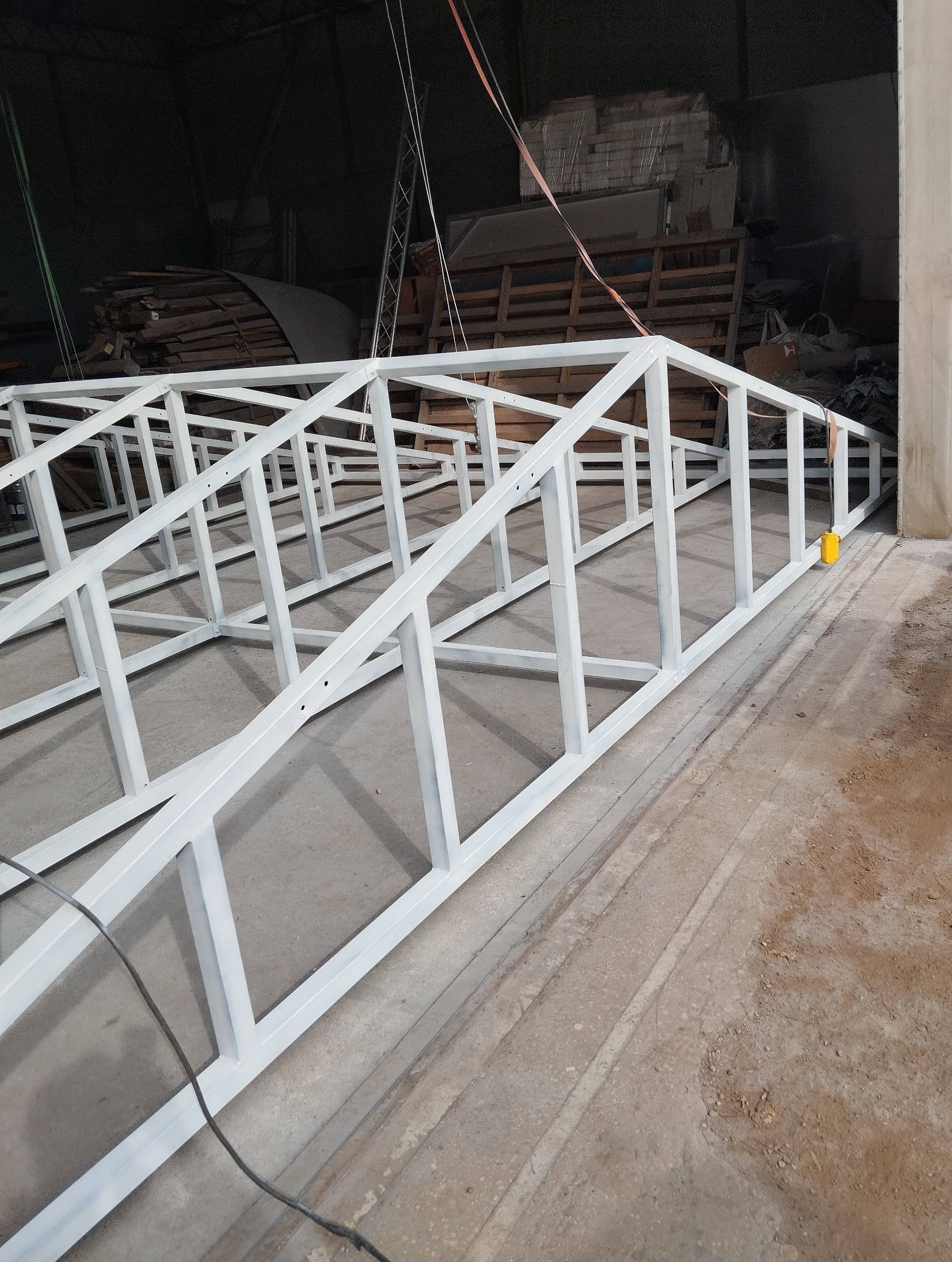 Biała, metalowa konstrukcja szkieletowa, prawdopodobnie brama lub ogrodzenie, leżąca na betonowej posadzce w hali produkcyjnej, widoczne ślady spawania i niedoskonałości powłoki malarskiej.