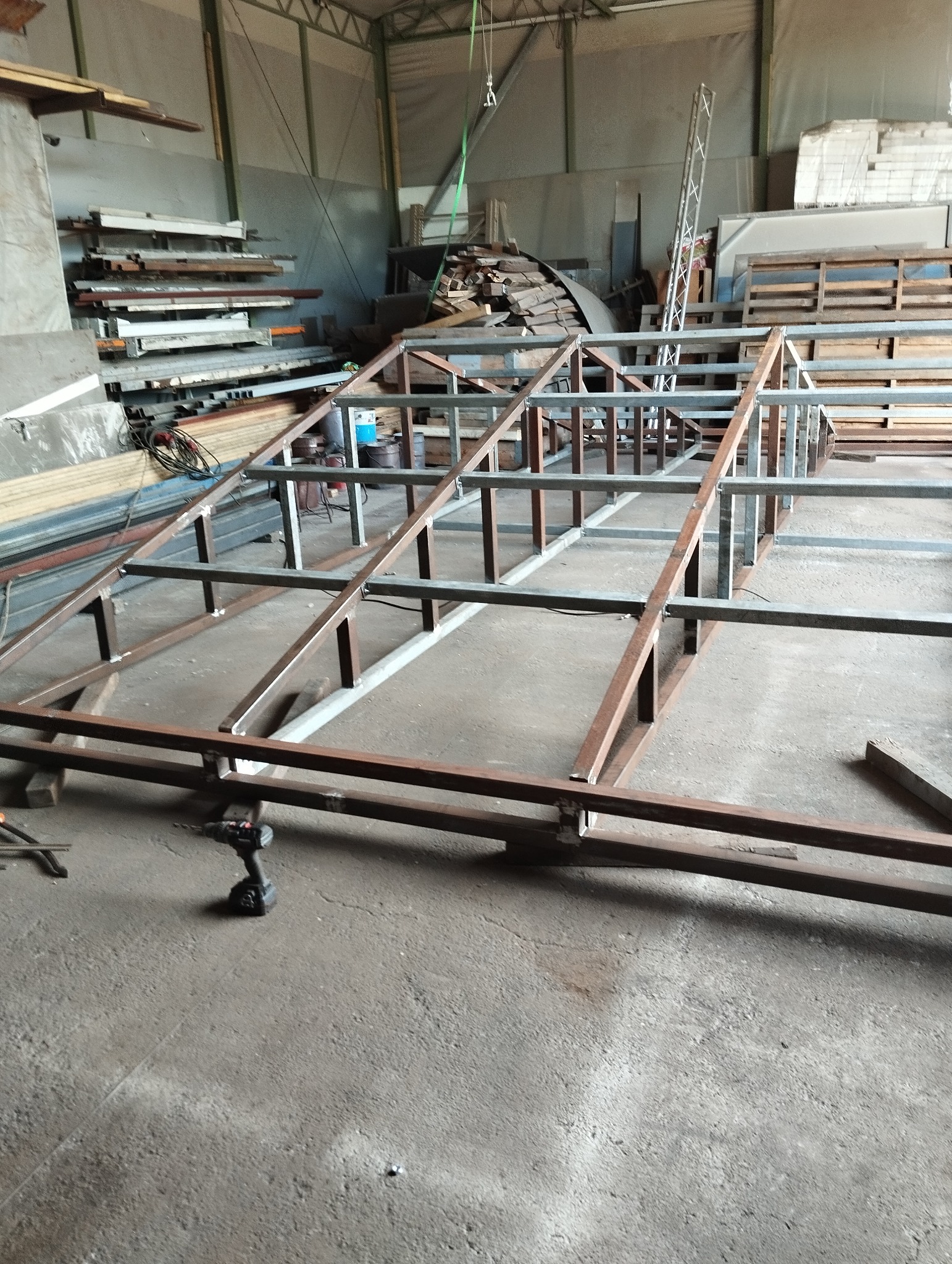 Stalowa konstrukcja szkieletowa, prawdopodobnie zespawana, leżąca na betonowej podłodze w warsztacie, obok wiertarko-wkrętarka akumulatorowa.