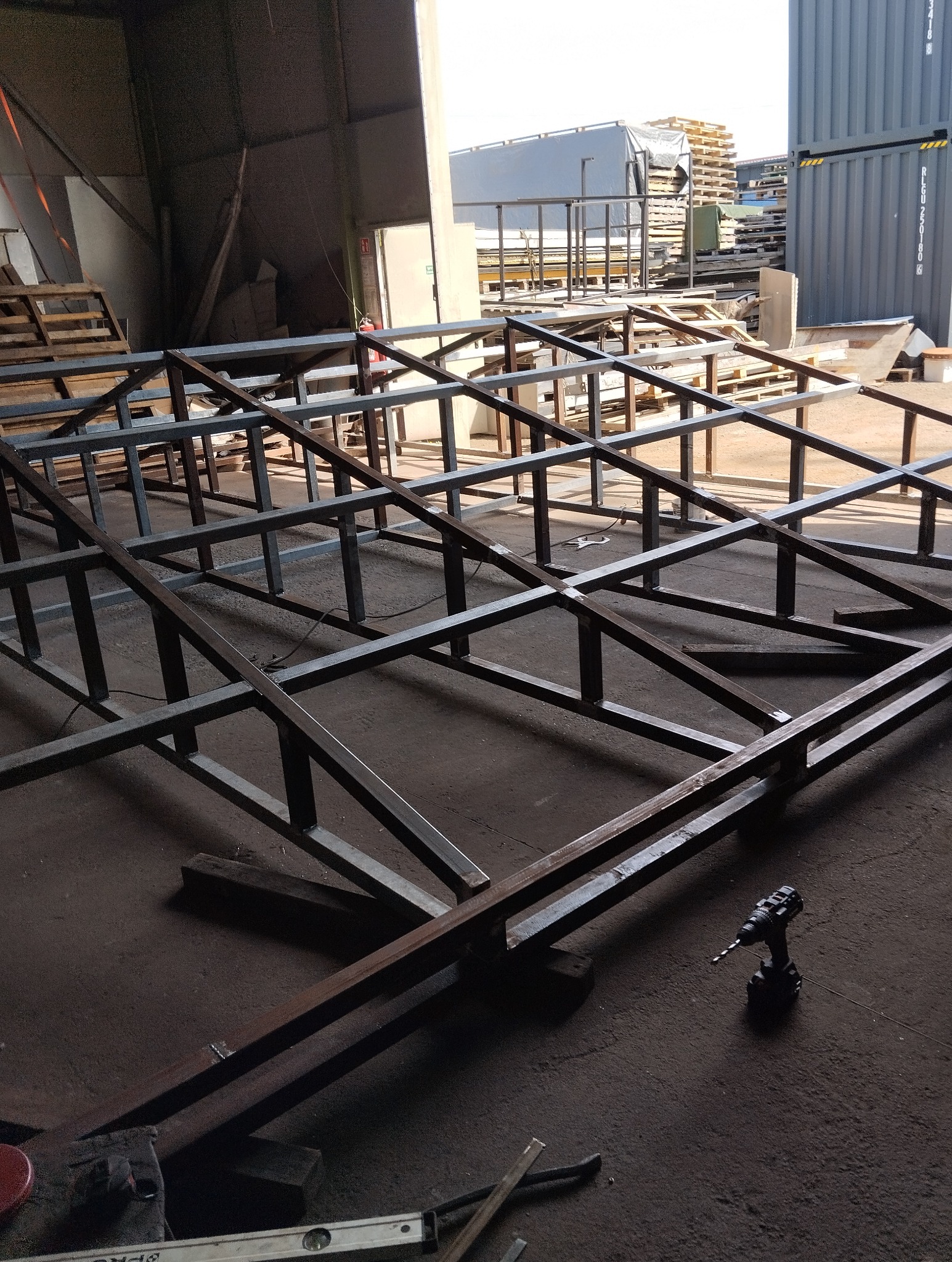 Konstrukcja szkieletowa z metalowych profili w hali produkcyjnej, widoczna wiertarka akumulatorowa, poziomica i fragmenty materiałów.