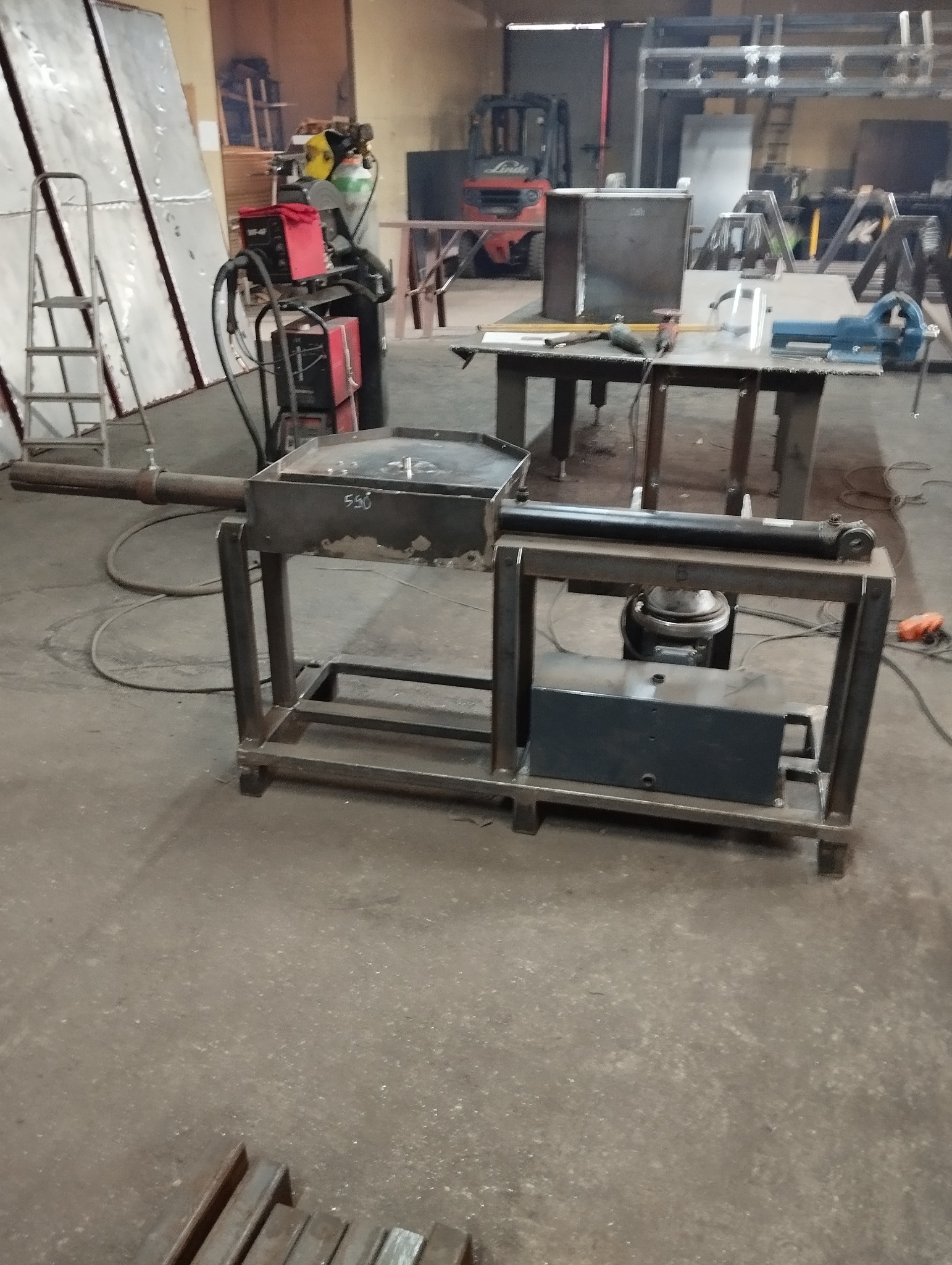 Konstrukcja metalowa z hydraulicznym siłownikiem i zbiornikiem, spawana z profili stalowych, widoczna w warsztacie, w tle spawarka, butle z gazem technicznym i wózek widłowy.