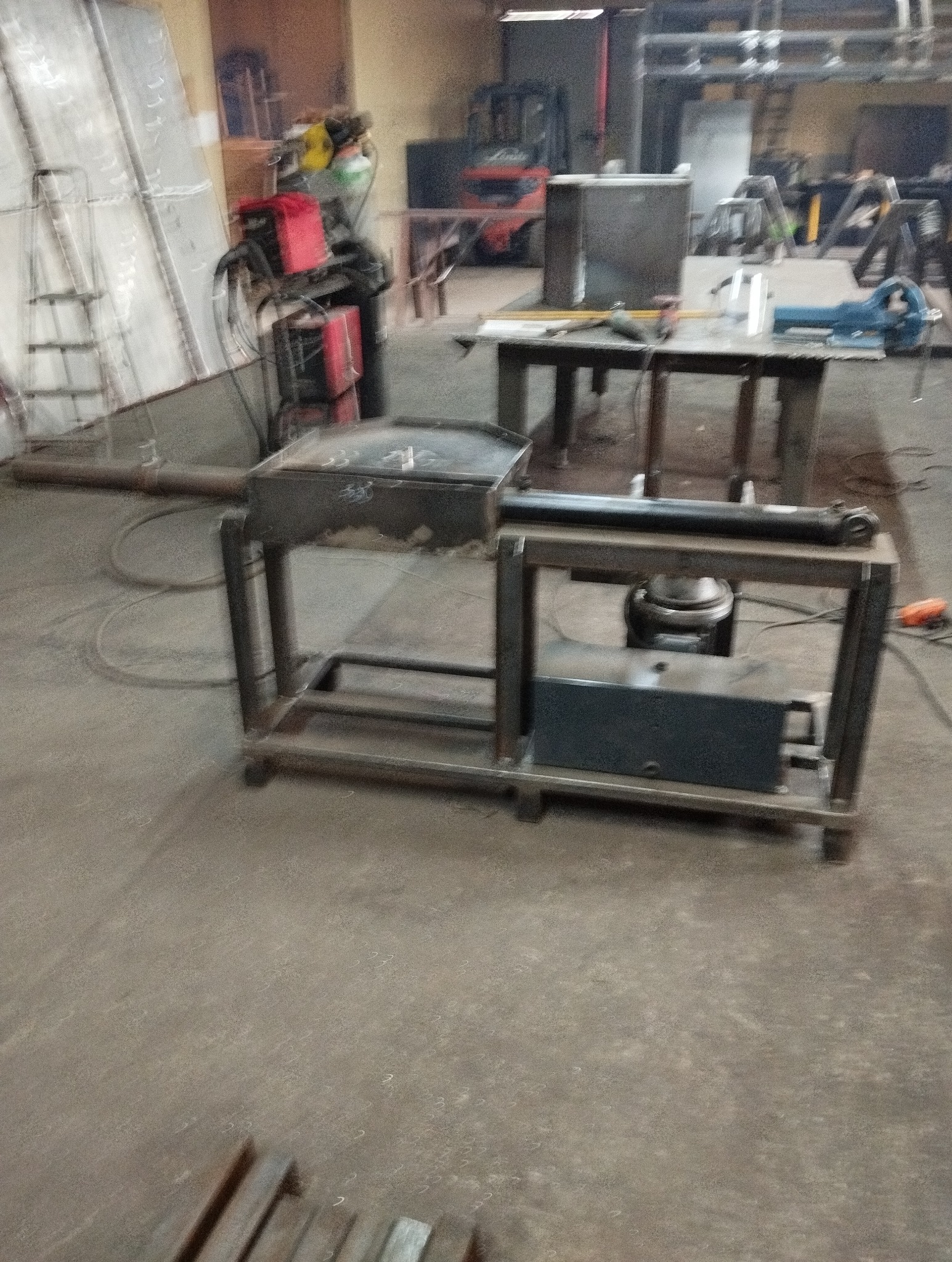 Warsztatowa sceneria z metalowym stołem roboczym, spawarką i wózkiem widłowym w tle, ukazująca środowisko pracy ślusarza lub spawacza.