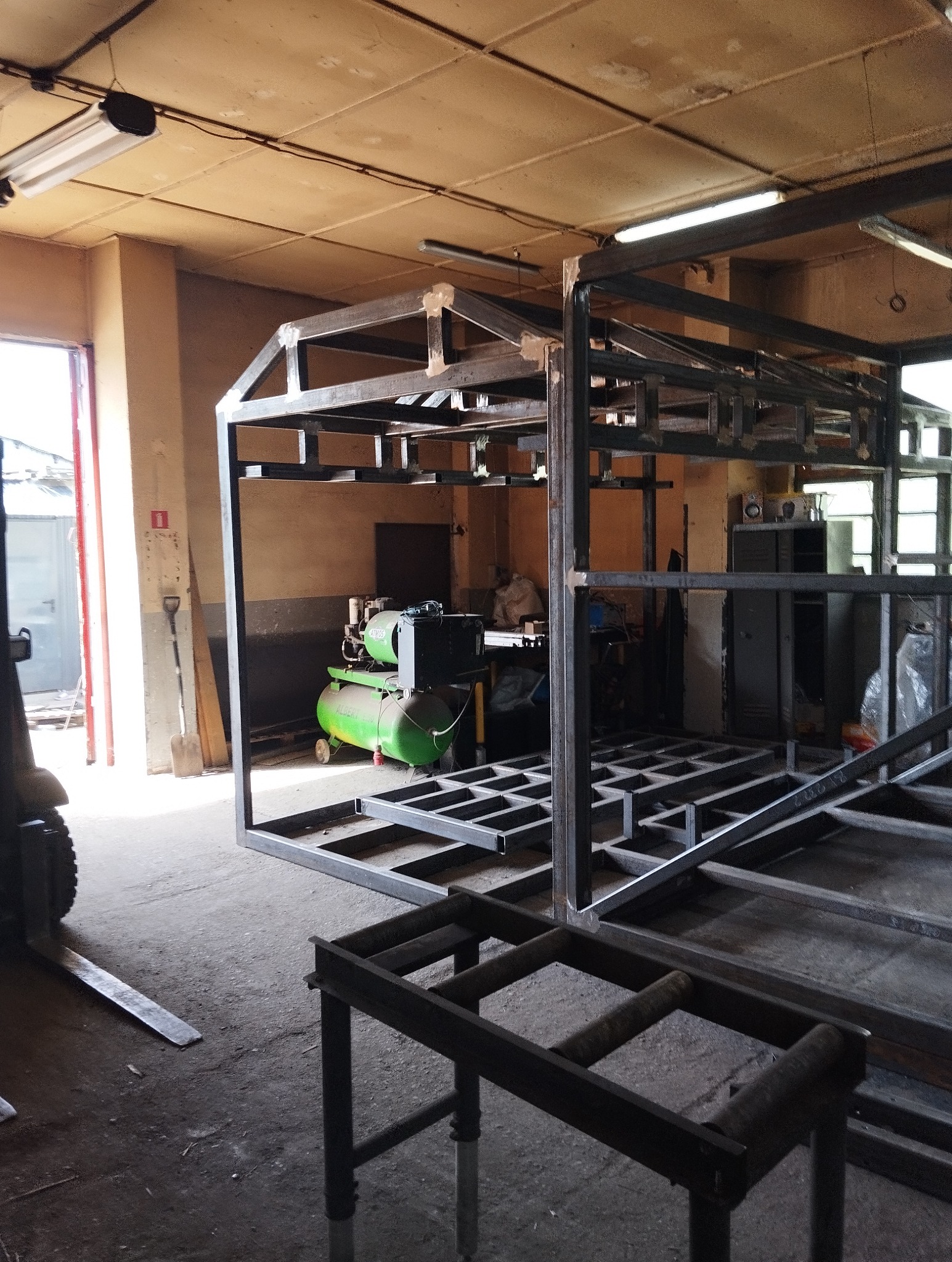 Konstrukcja szkieletowa z metalowych profili, spawana w warsztacie, widoczny kompresor i wózek widłowy w tle.