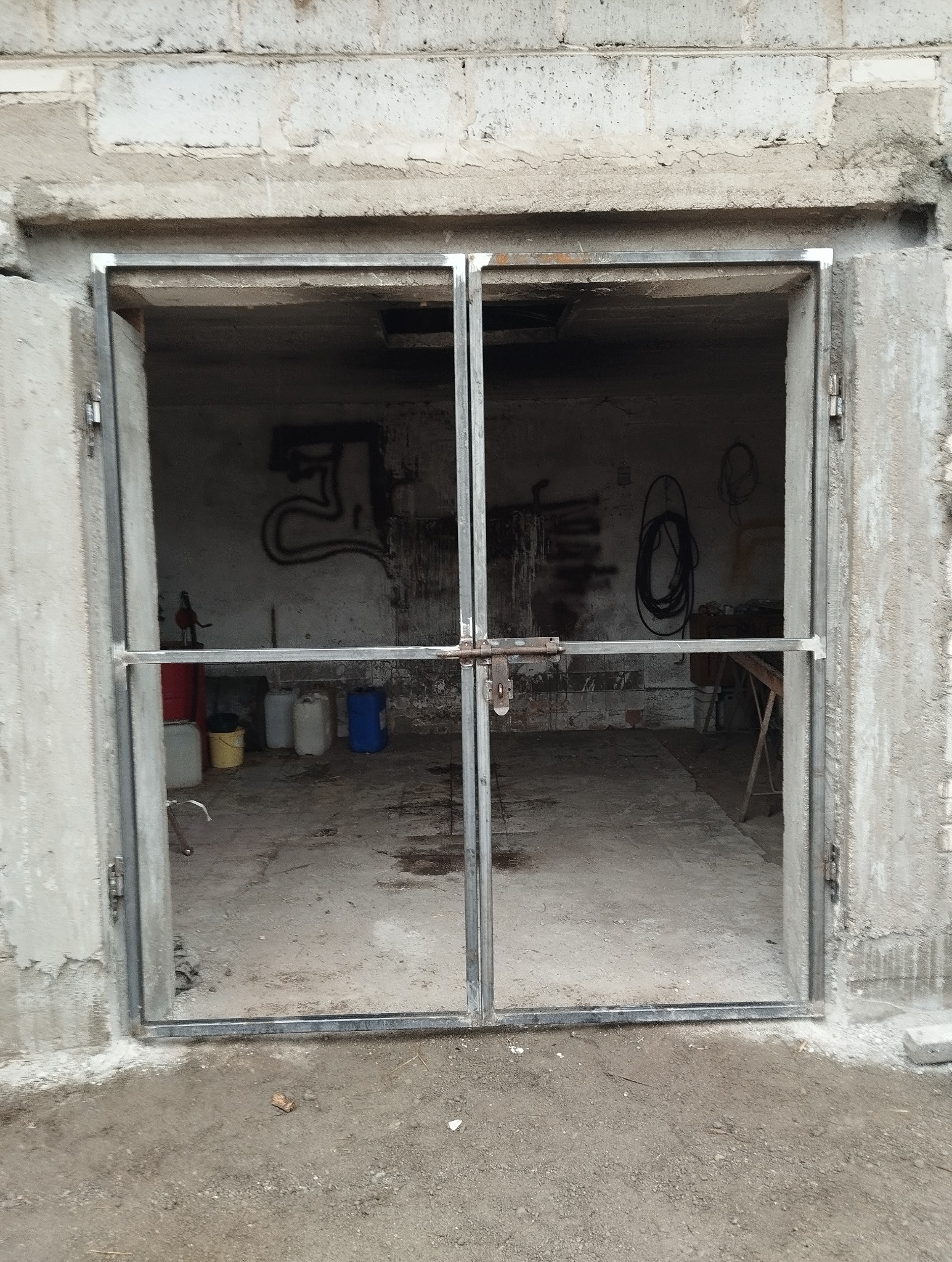 Metalowa rama dwuskrzydłowej bramy w surowym stanie, zamontowana w otworze budynku z betonowych pustaków, widoczny zamek na środku, wnętrze pomieszczenia z różnymi przedmiotami w tle.