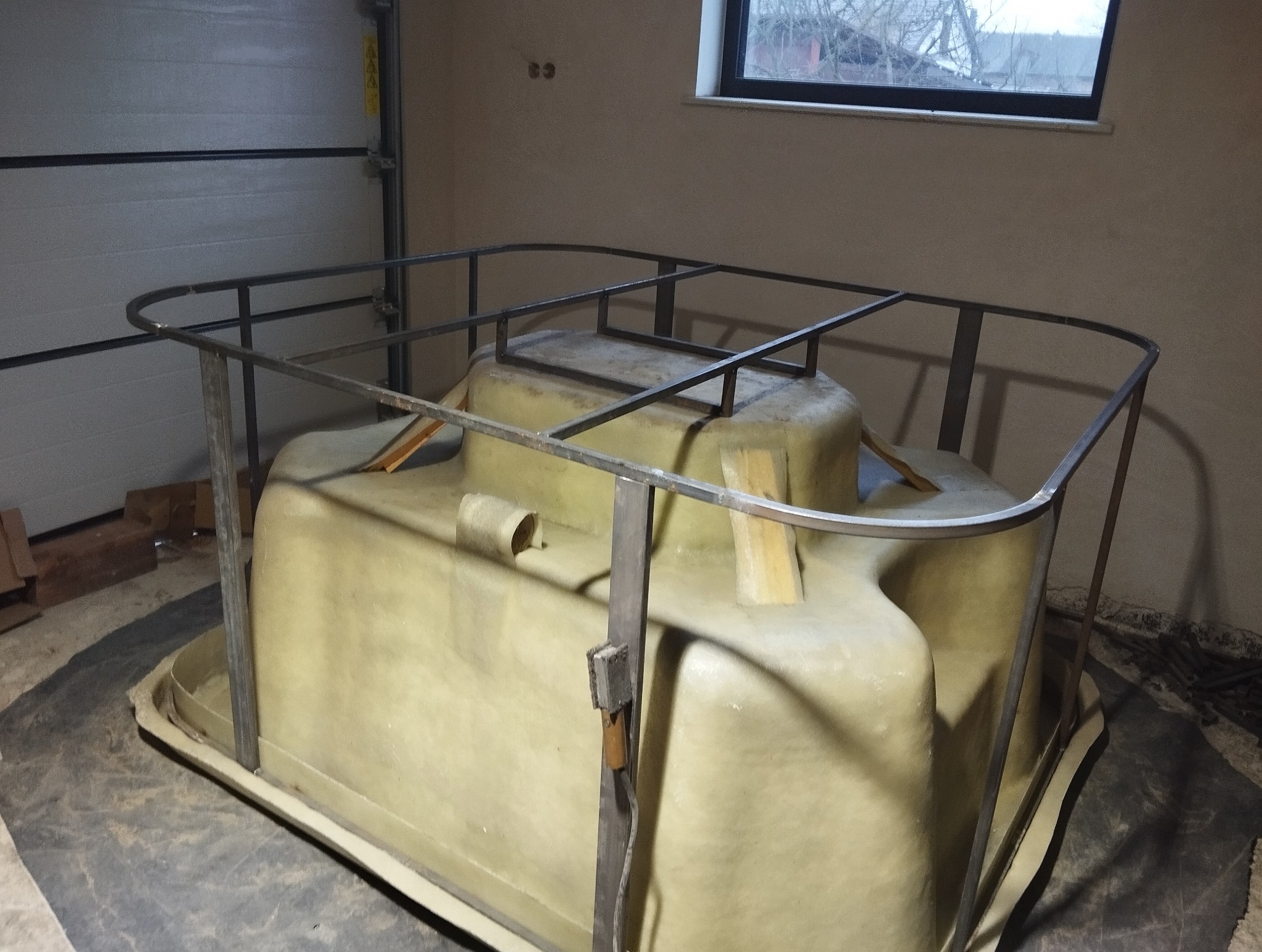 Kanciasta, beżowa forma z tworzywa sztucznego w garażu, zabezpieczona metalową ramą z profili kwadratowych, widoczne drewniane kliny i spawarka punktowa.