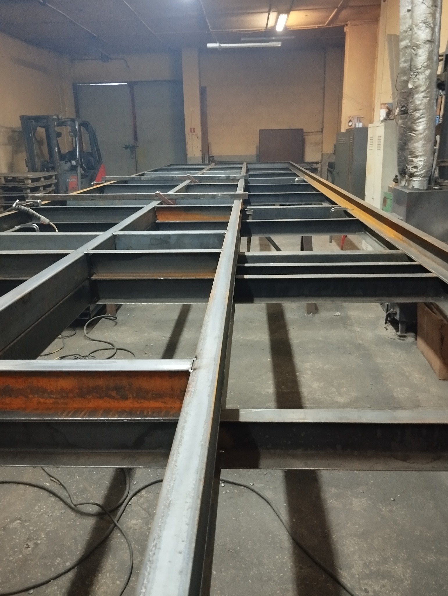 Konstrukcja stalowa z profili zamkniętych w warsztacie, widoczne spawy i przewody spawalnicze na podłodze, w tle wózek widłowy.