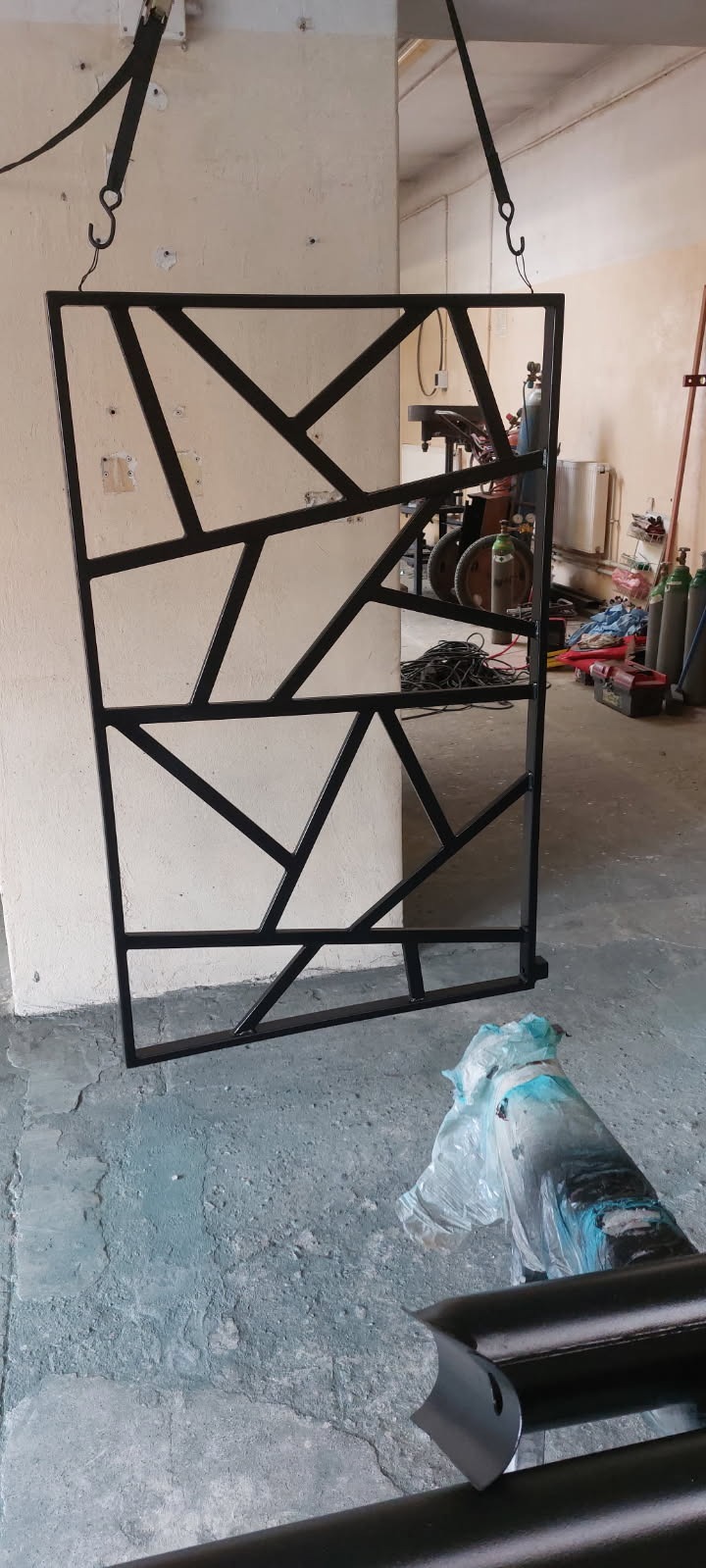 Metalowa konstrukcja z geometrycznym wzorem, zawieszona na hakach w warsztacie, z widocznymi narzędziami i butlami gazowymi w tle.