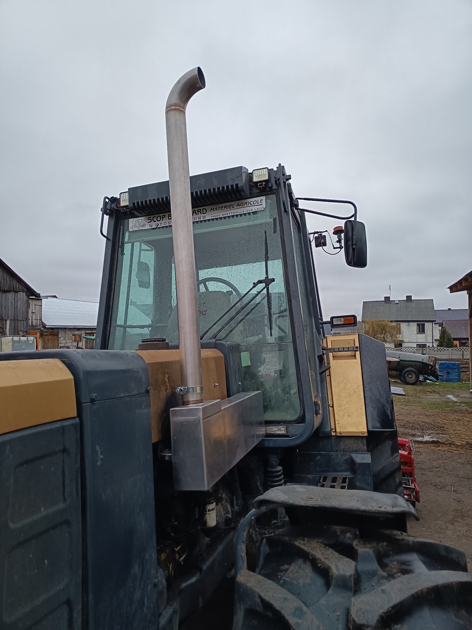 Spawana rura wydechowa ze stali nierdzewnej zamontowana na ciągniku rolniczym, widok z tyłu kabiny, widoczne spawy i mocowania, na tle zabudowania gospodarcze i pochmurne niebo.