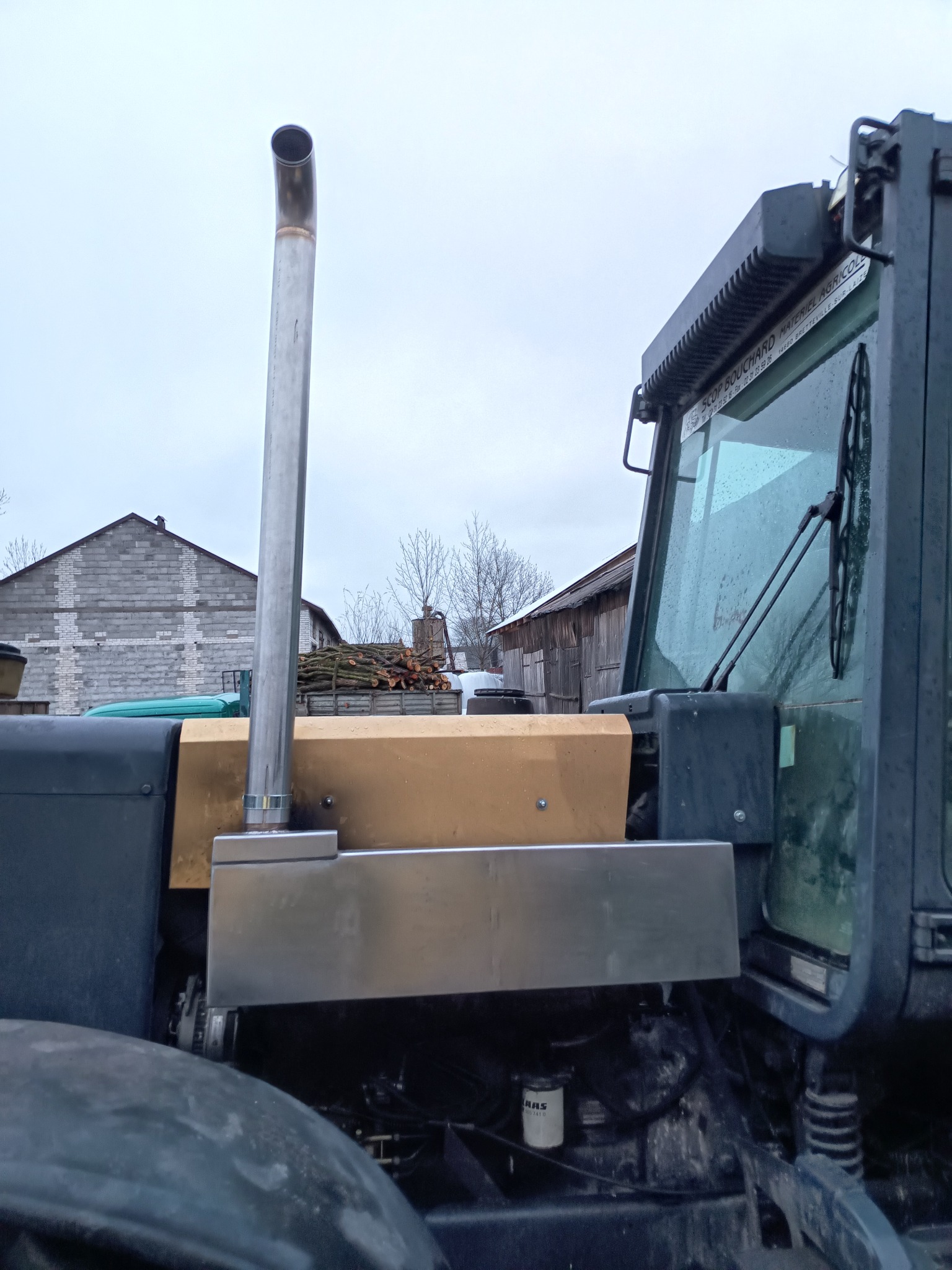 Fragment ciągnika rolniczego z widocznym kominem spalinowym ze stali nierdzewnej, fragmentem kabiny i silnikiem, na tle budynków gospodarczych i pochmurnego nieba.