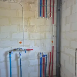 Instalacja wodna i kanalizacyjna w surowym wnętrzu: piony rur w izolacji (czerwone i niebieskie), szara rura kanalizacyjna, białe przewody elektryczne przy ścianie z betonu komórkowego.