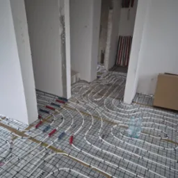 Białe rury ogrzewania podłogowego ułożone na czarnej siatce na folii aluminiowej. W tle widoczne piony instalacji wodnej i surowe ściany w trakcie remontu.