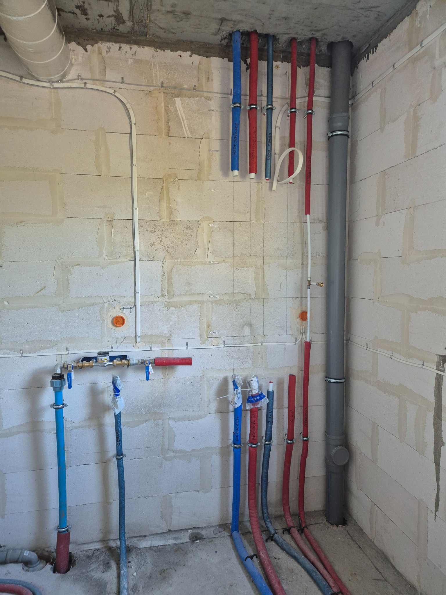Instalacja wodna i kanalizacyjna w surowym wnętrzu: piony rur w izolacji (czerwone i niebieskie), szara rura kanalizacyjna, białe przewody elektryczne przy ścianie z betonu komórkowego.