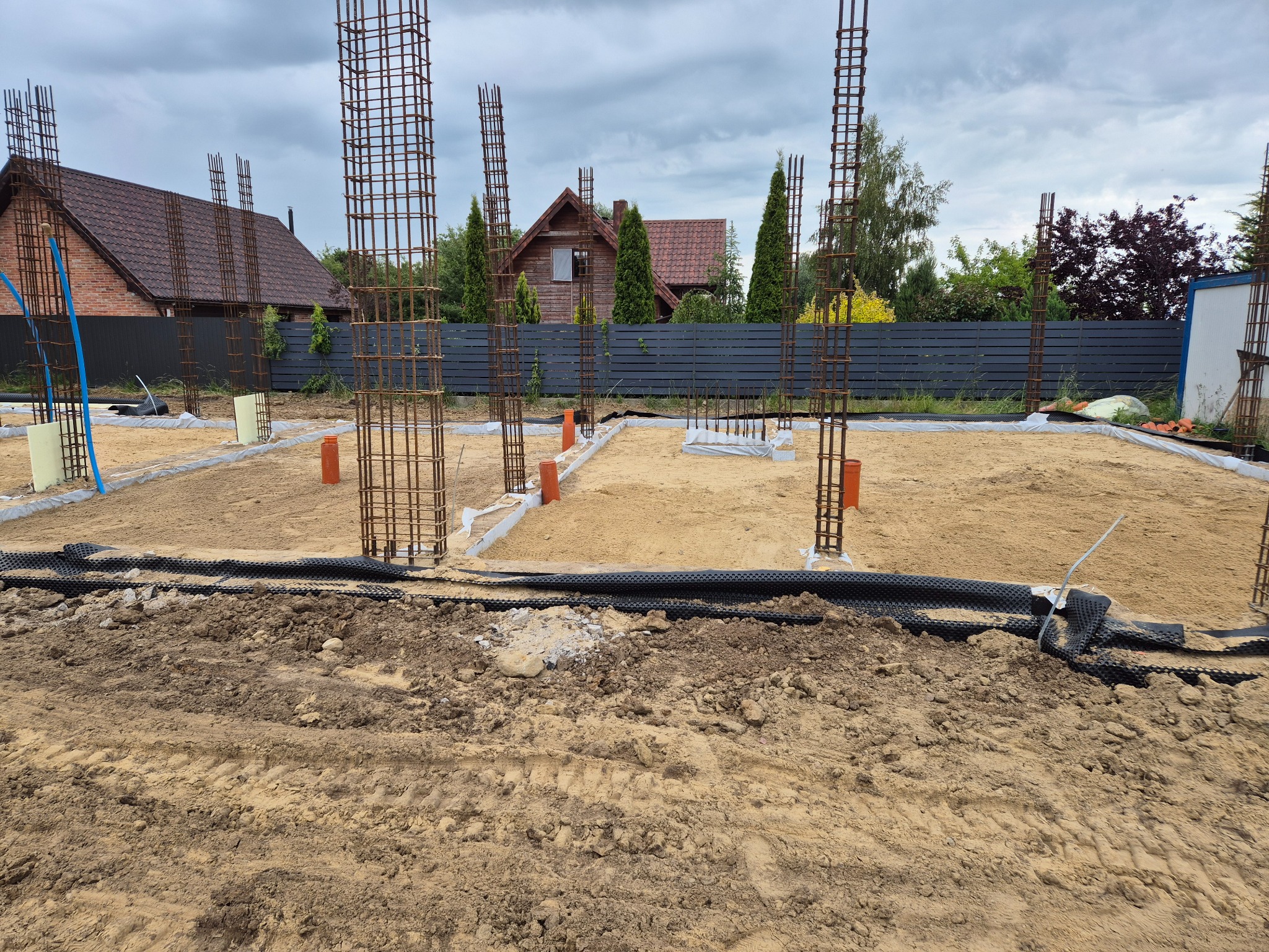 Fundamenty domu w trakcie budowy: zbrojenie, izolacja, grunt. Widoczne elementy konstrukcyjne i przygotowanie terenu pod wylewkę. W tle zabudowa jednorodzinna i pochmurne niebo.