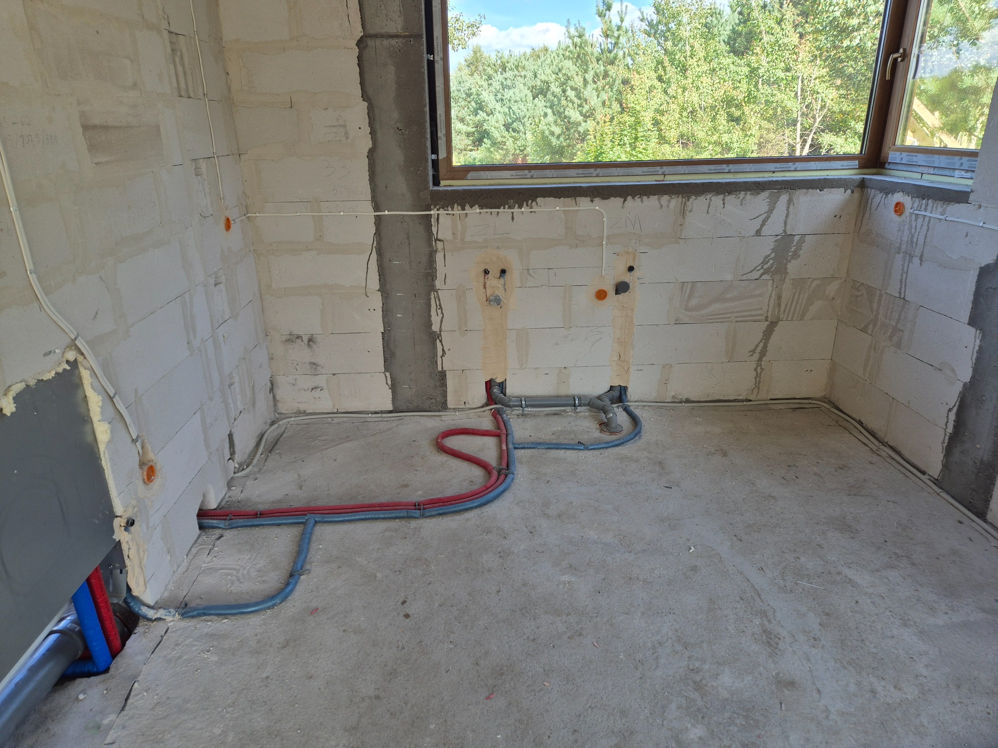 Surowy stan: instalacja hydrauliczna i elektryczna w ścianach z betonu komórkowego, widok na okno z zielenią. Czerwone i niebieskie rury na podłodze, pomarańczowe puszki elektryczne.
