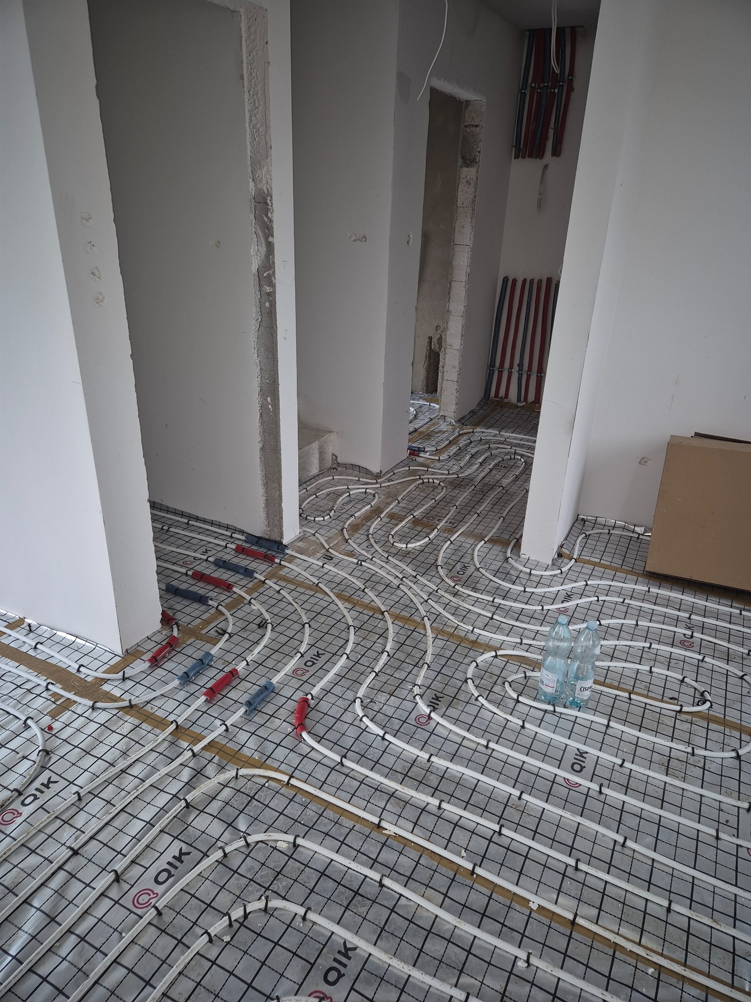 Białe rury ogrzewania podłogowego ułożone na czarnej siatce na folii aluminiowej. W tle widoczne piony instalacji wodnej i surowe ściany w trakcie remontu.
