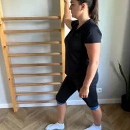 Kobieta w sportowym stroju rozciąga boczną część tułowia, opierając się o drabinki gimnastyczne w jasnym wnętrzu.