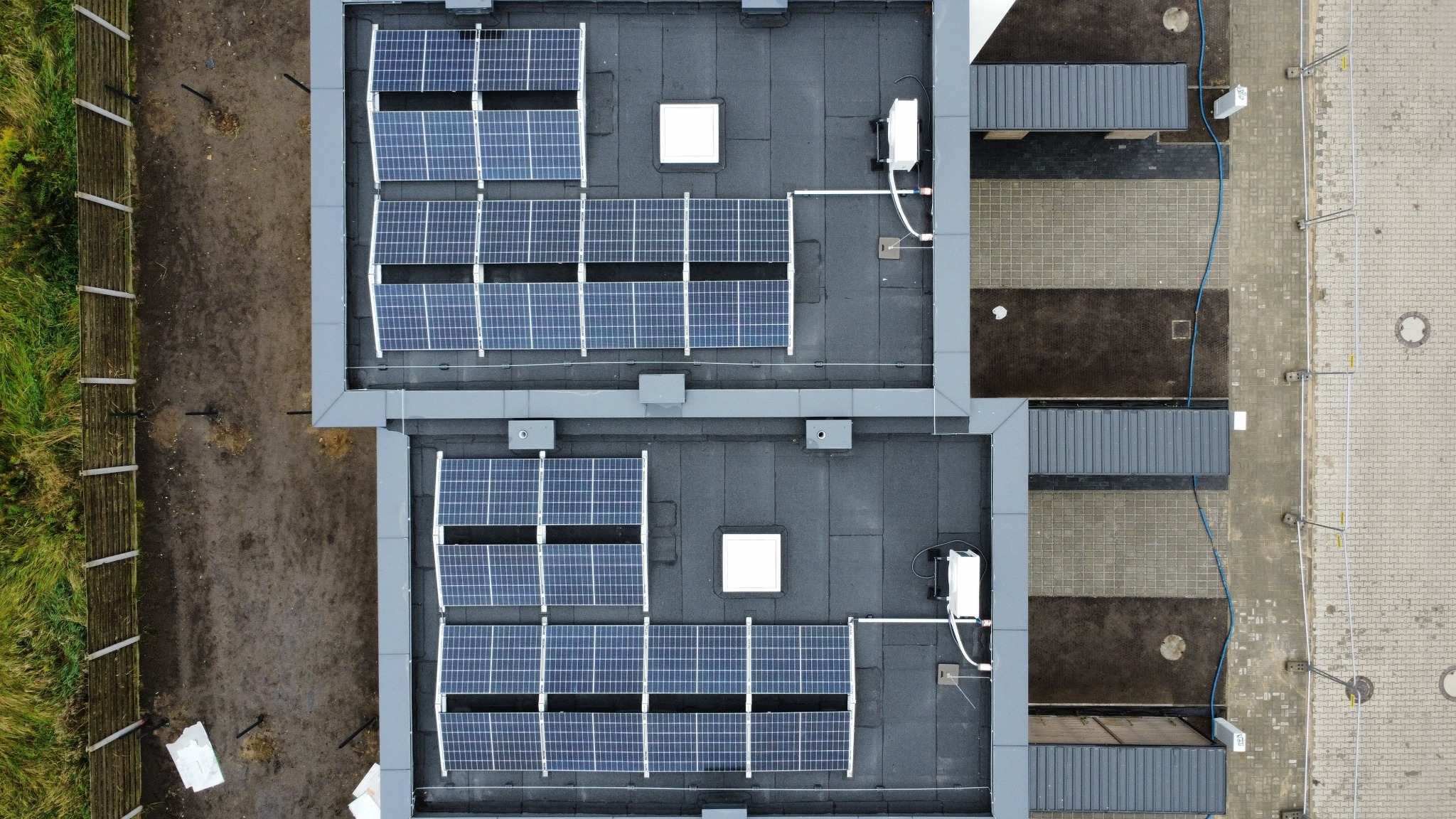 Widok z góry na dwa dachy budynków z zainstalowanymi panelami słonecznymi, białe falowniki przy krawędzi dachu, obok budynków kostka brukowa i drewniany płot.