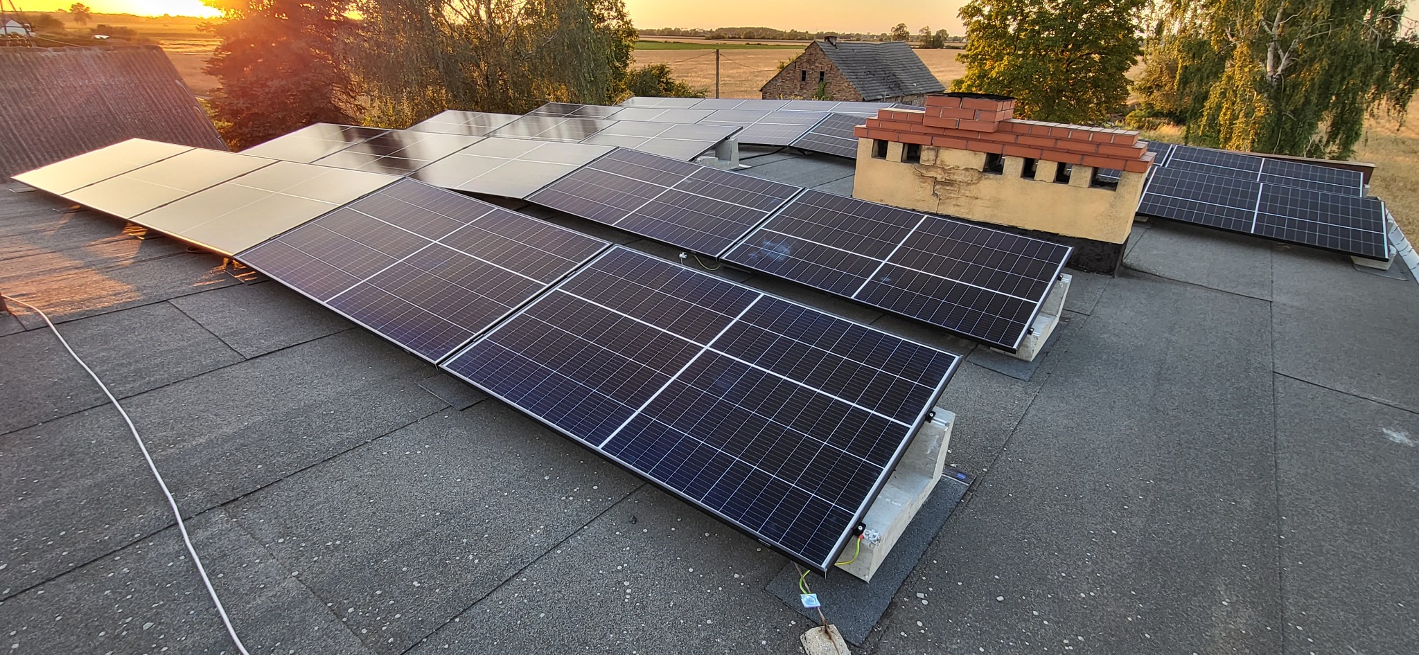 Instalacja fotowoltaiczna na dachu z papy, panele zamontowane na betonowych podstawach, w tle budynek gospodarczy i zachodzące słońce.